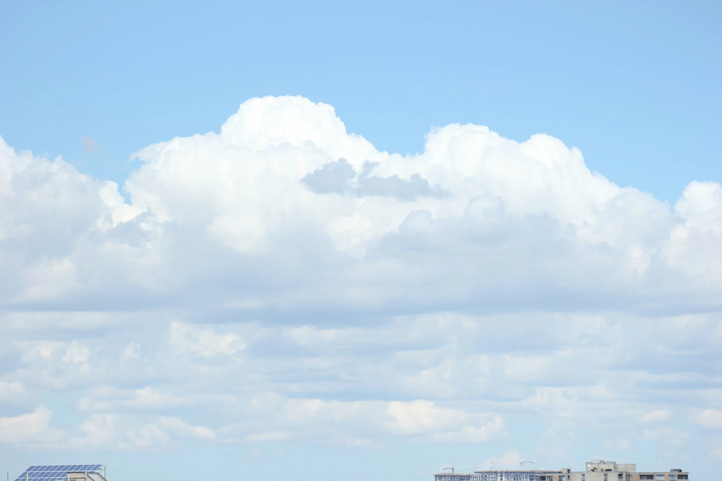 Clear blue sky with large white fluffy clouds, and a cityscape with buildings and solar panels at the bottom.