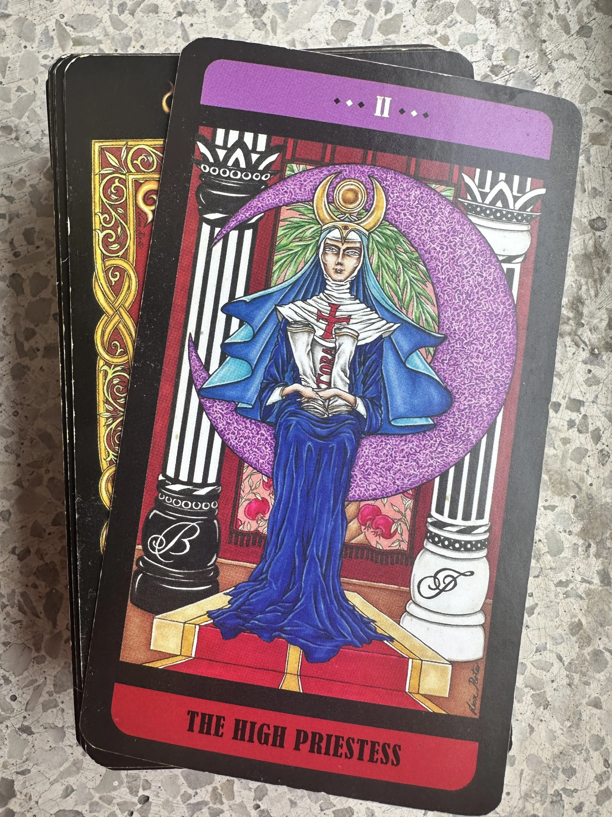 Tarot card titled 'The High Priestess' featuring a figure dressed in blue robes with a crown and cloak, seated on a throne with pillars decorated with initials B and D, set against a detailed background with purple, red, and white colors.