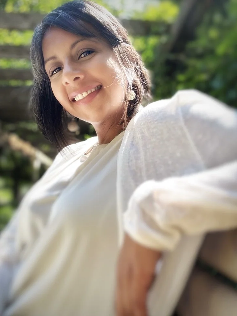 A woman smiling outdoors with sunlight on her face, wearing a white top and earrings, with green foliage in the background.