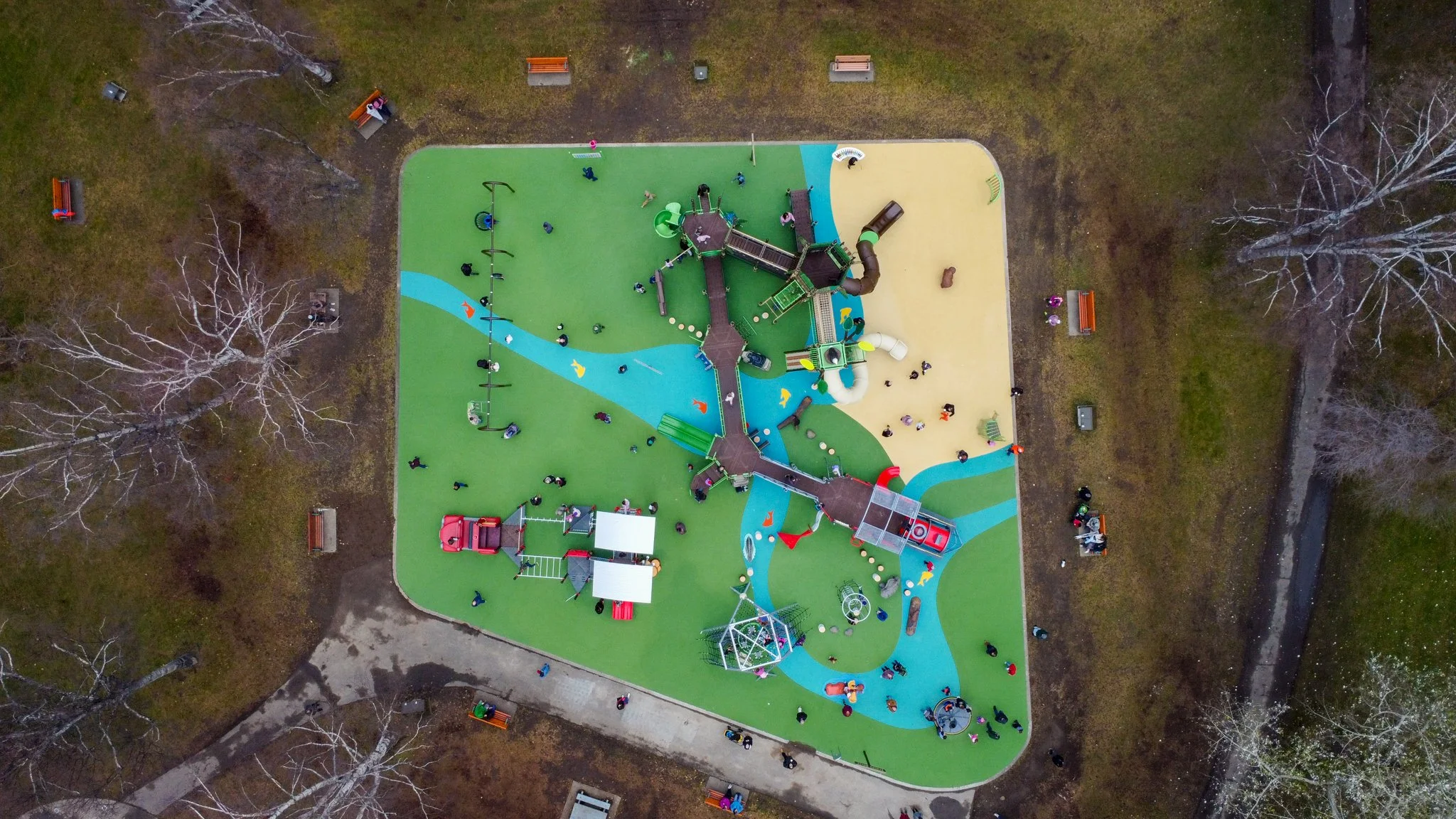 A top-down aerial view of a colorful playground with various play structures, slides, and swings surrounded by trees and benches.
