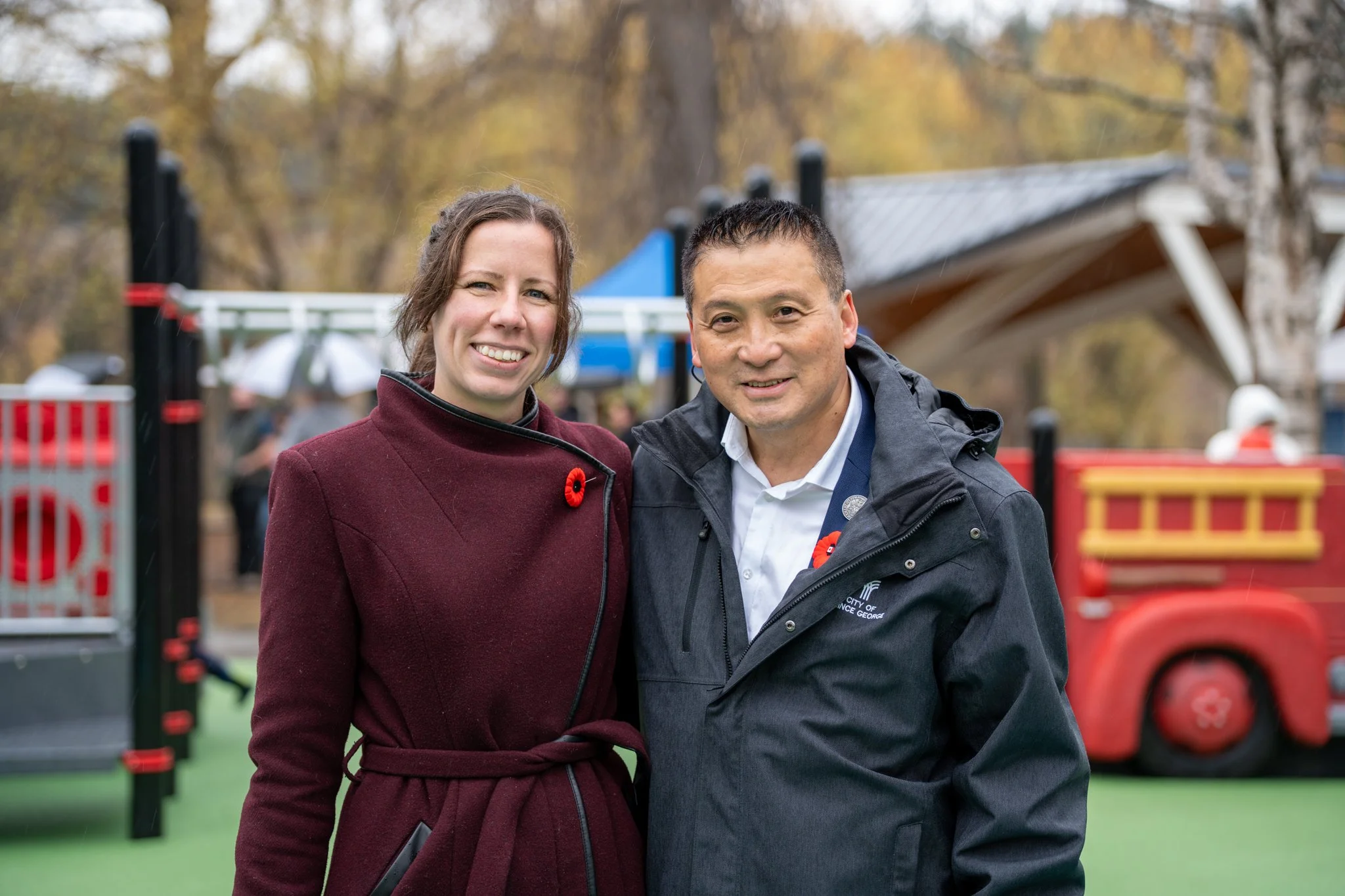 Two people smiling outdoors, a woman wearing a maroon coat and a man in a black jacket, with a playground and trees in the background.