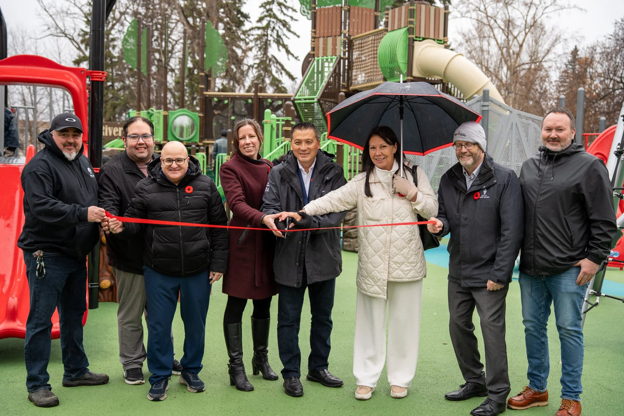 Group of eight people holding a red ribbon at a playground opening, with some holding a pair of scissors, under an umbrella, and smiling.