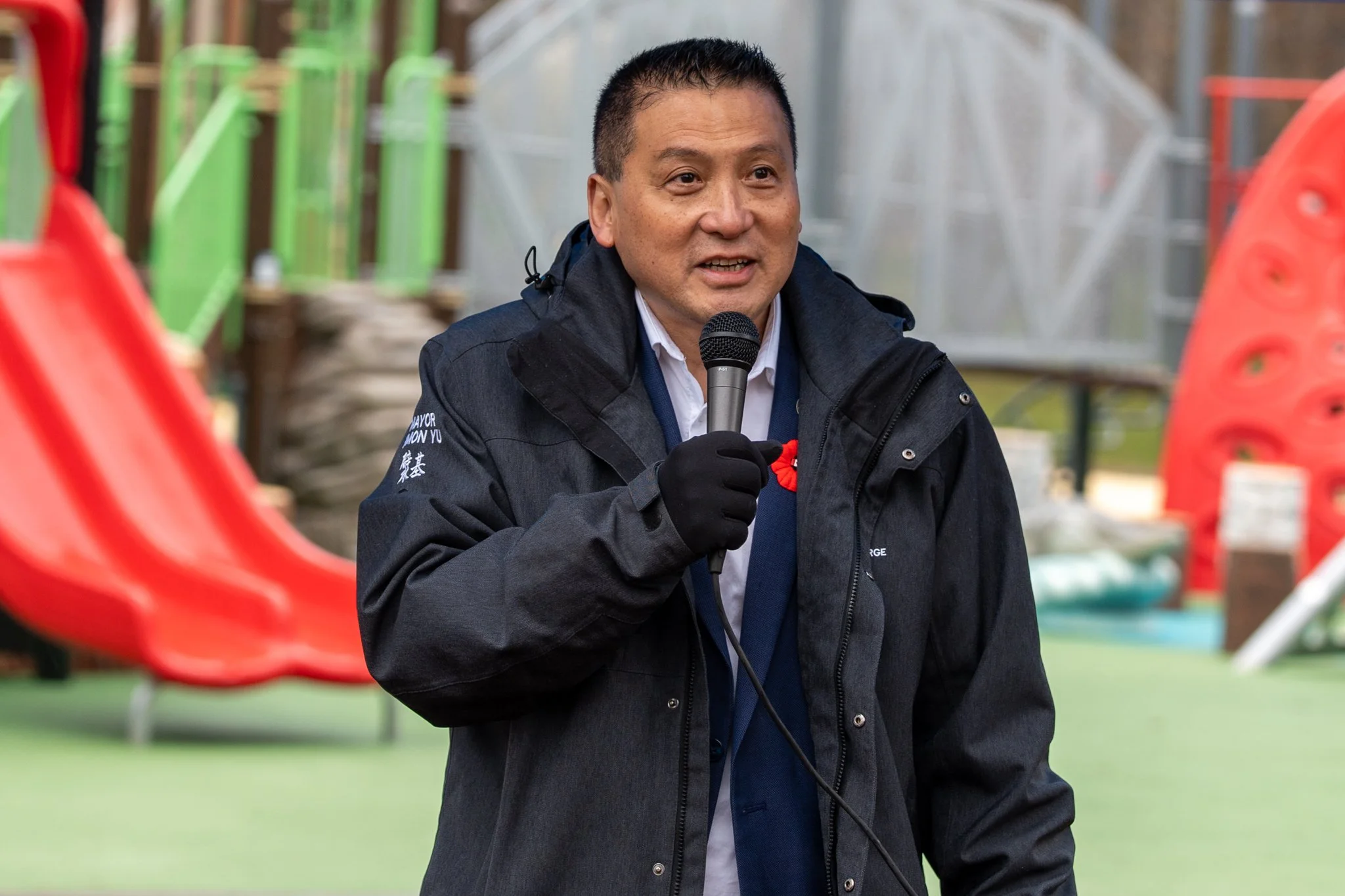 A man holding a microphone speaking outdoors, wearing a black jacket, with playground equipment in the background.