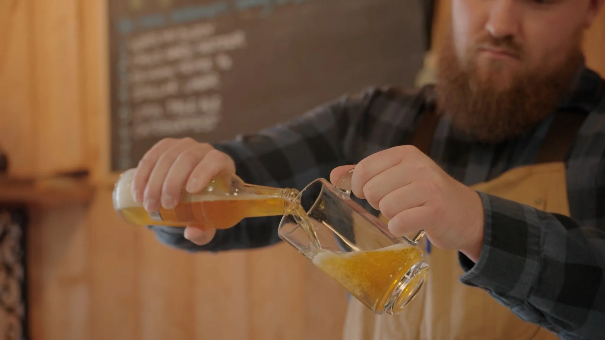 A man with a beard pouring beer into two glasses at a bar or brewery.