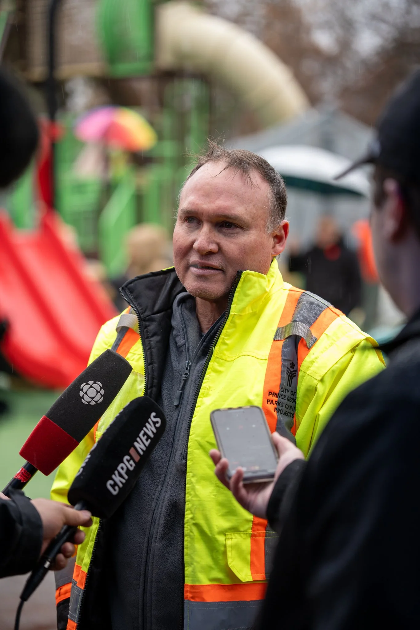 Man in a yellow safety jacket speaking to reporters with microphones and a person holding a smartphone in front of him outdoors.