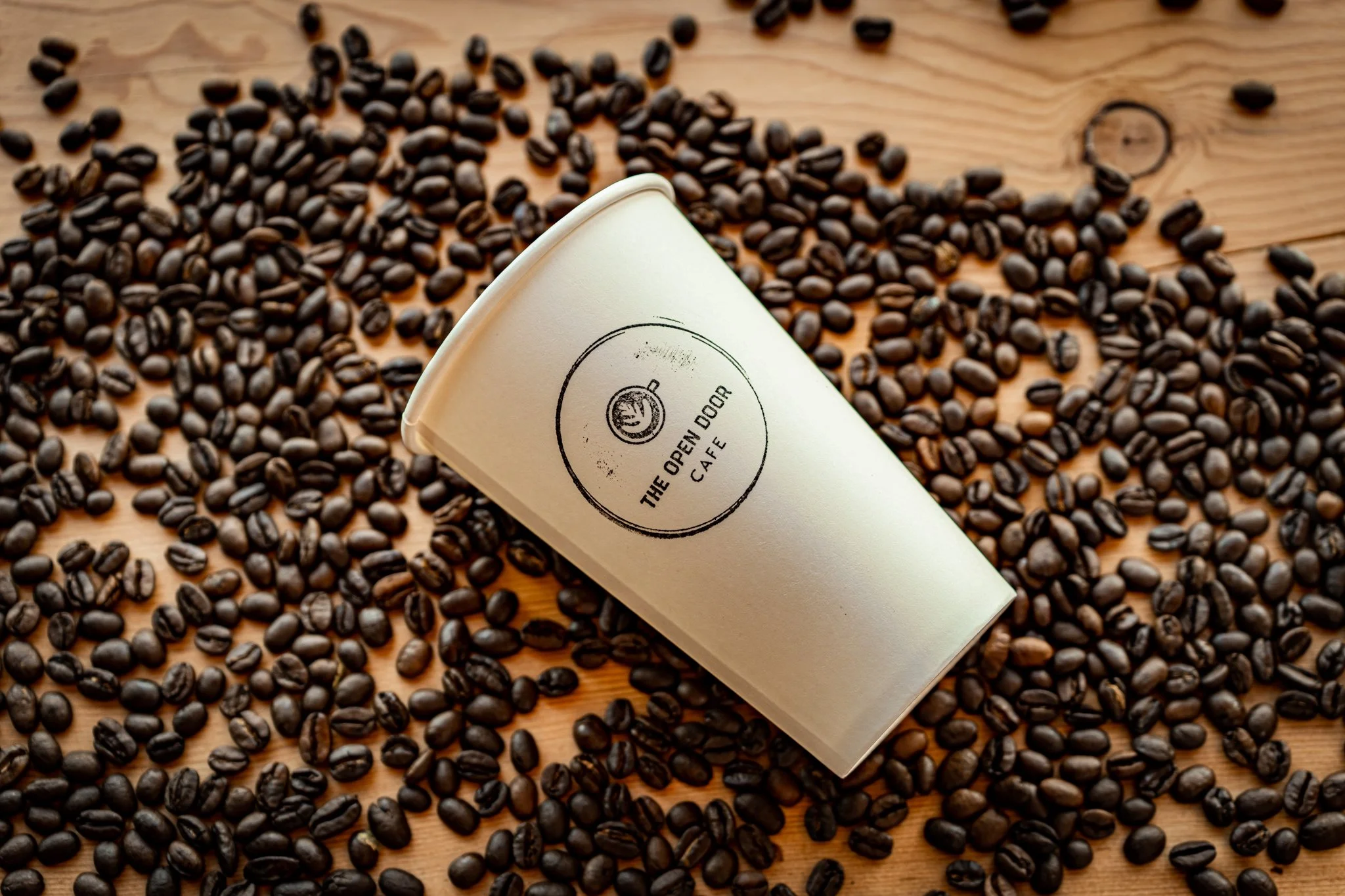 A coffee cup labeled 'The Open Door Cafe' resting on a wooden surface surrounded by scattered coffee beans.