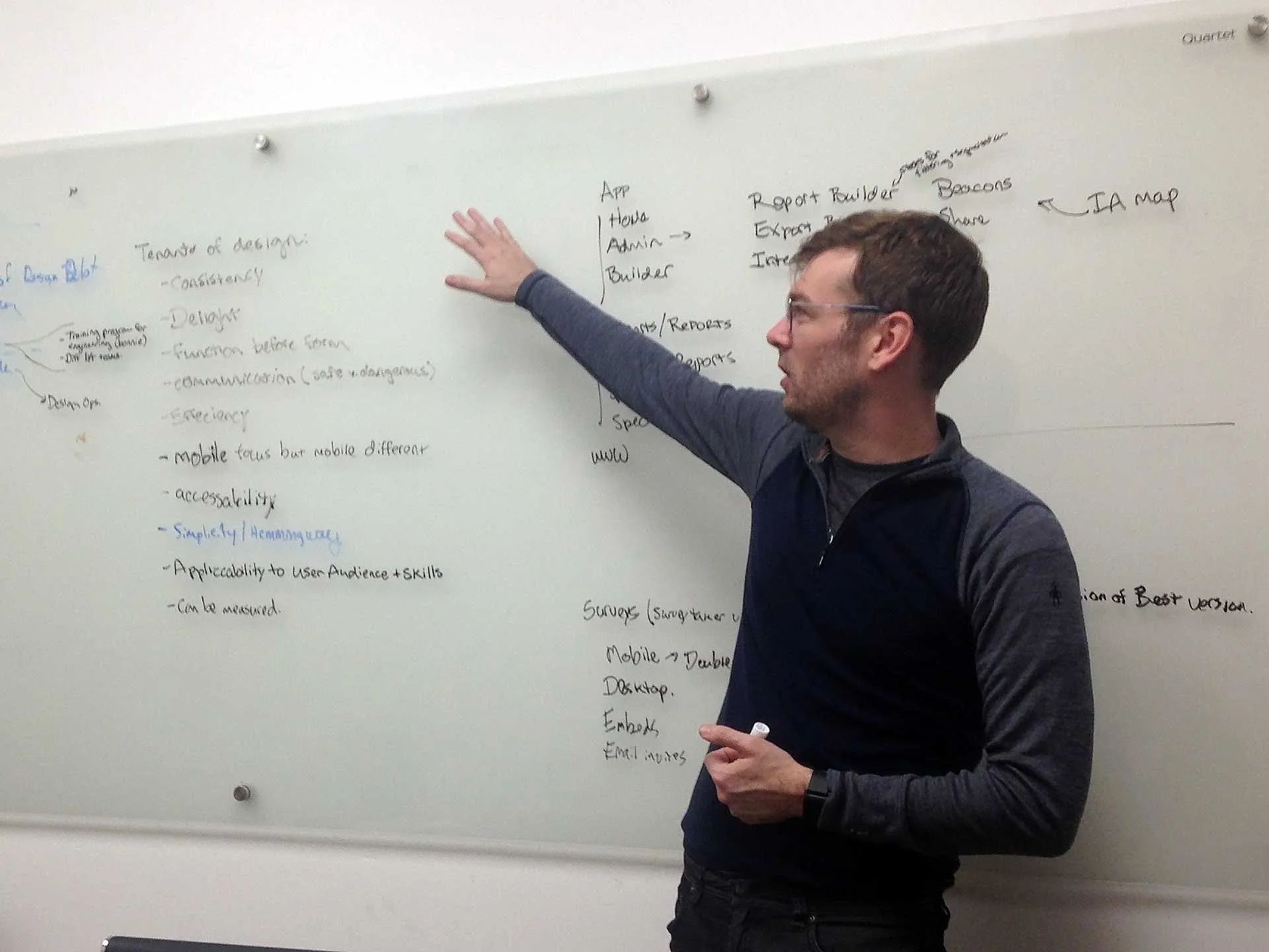 One designer, in front of a white board, holding a market and pointing at a written down list of requirements.