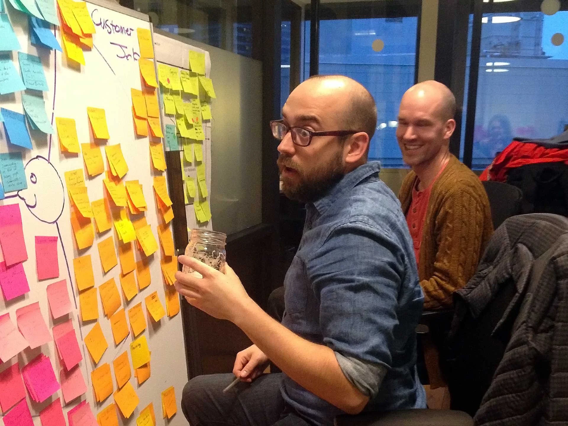 Two designers organizing findings from a user research study, in front of a white board with post-it notes.  In the window behind them it is getting dark.