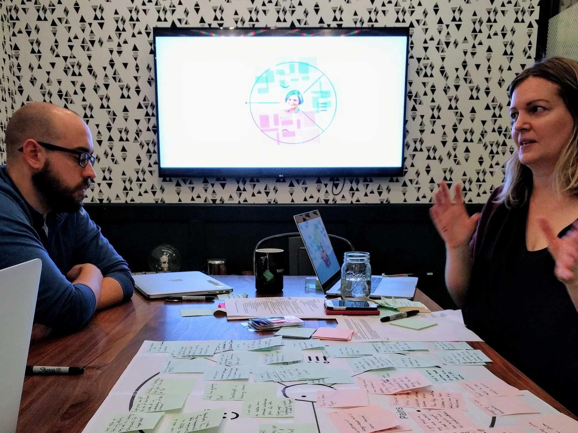 Two designers at a table with a post it notes and sheets of paper in front of them, animatedly talking about findings from a user research workshop.