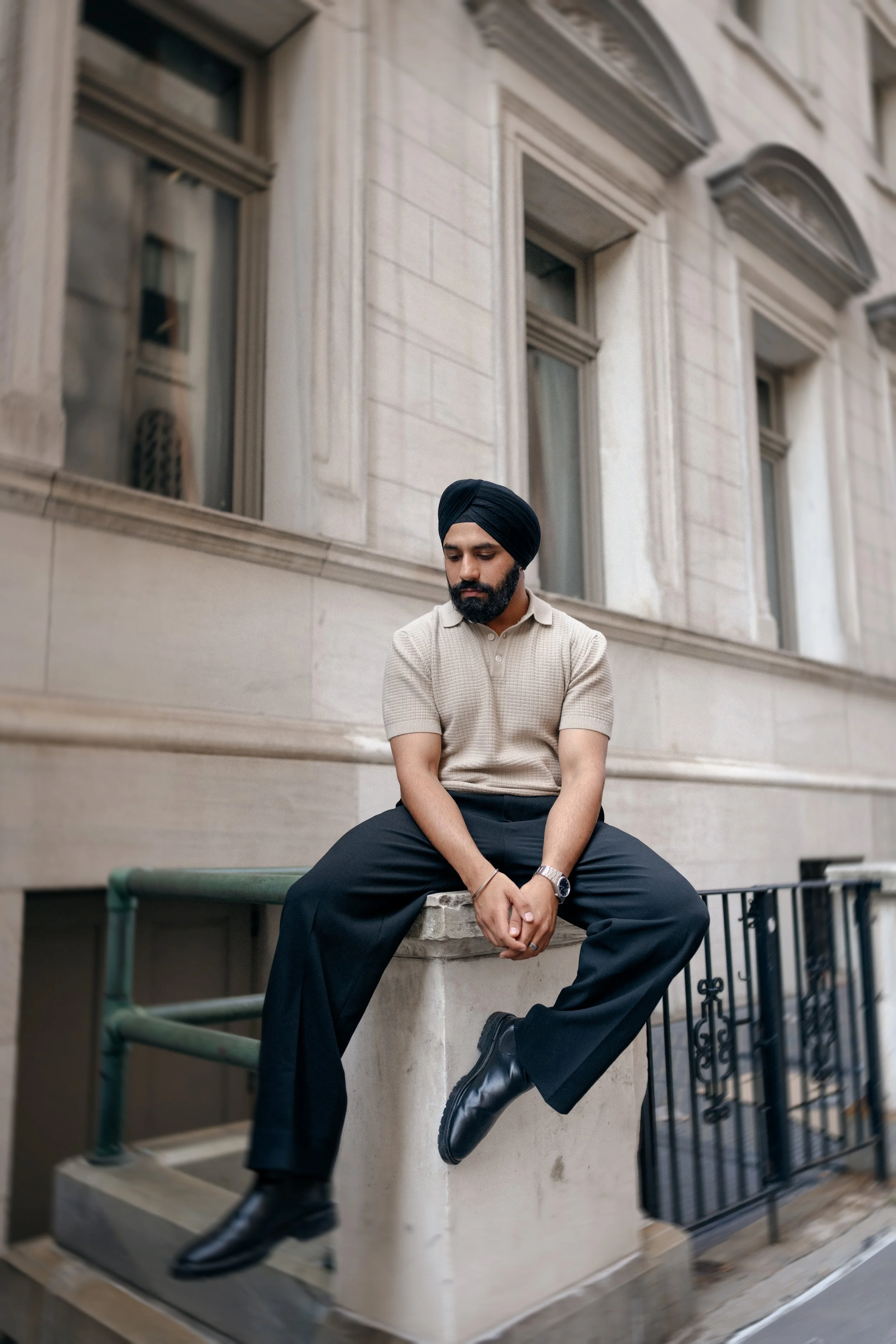 A man wearing a black turban, beige shirt, black pants, and black shoes sitting on a stone ledge outside a building with large windows and decorative architectural details.