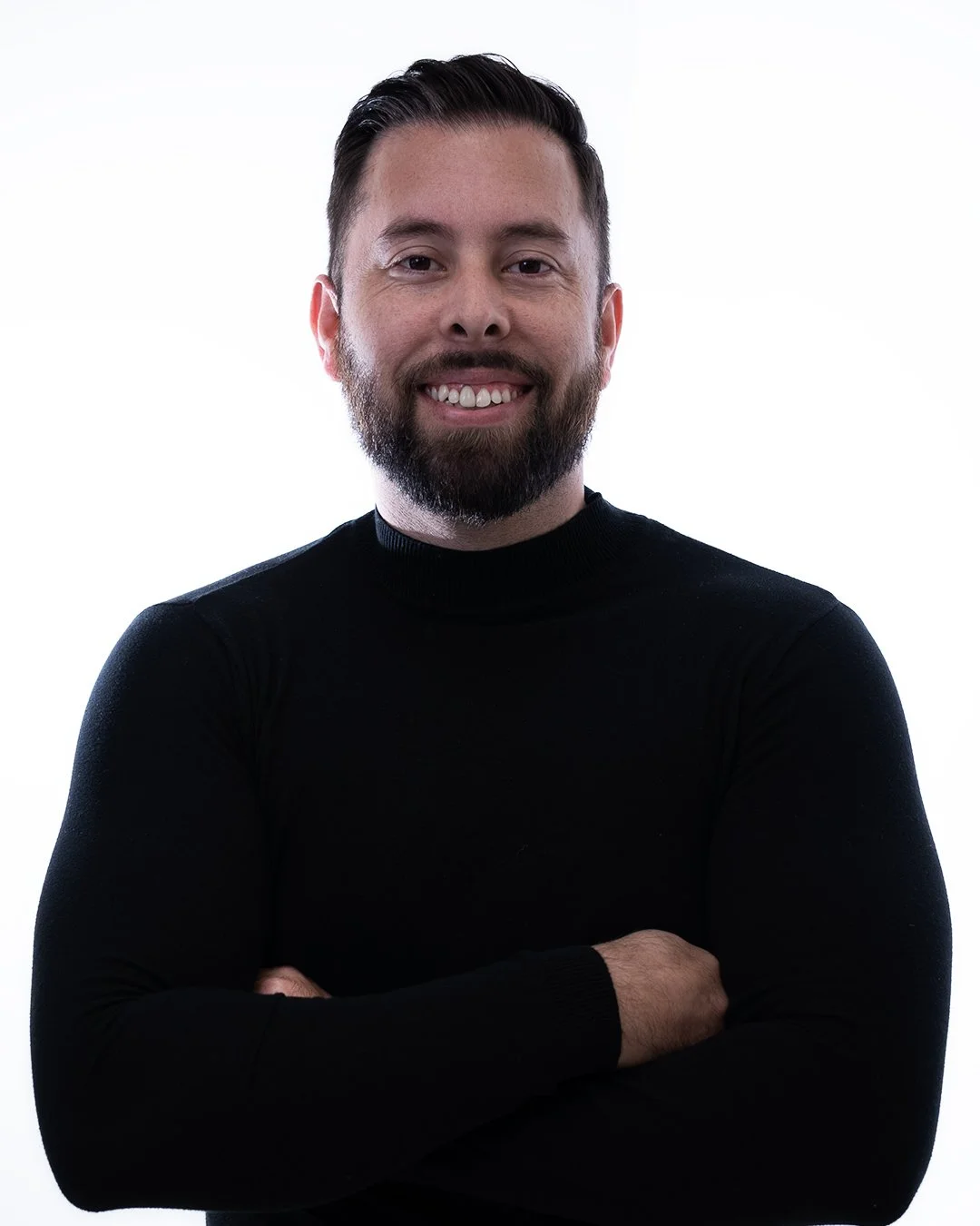 A man with dark hair and a beard wearing a black turtleneck, smiling with arms crossed against a white background.