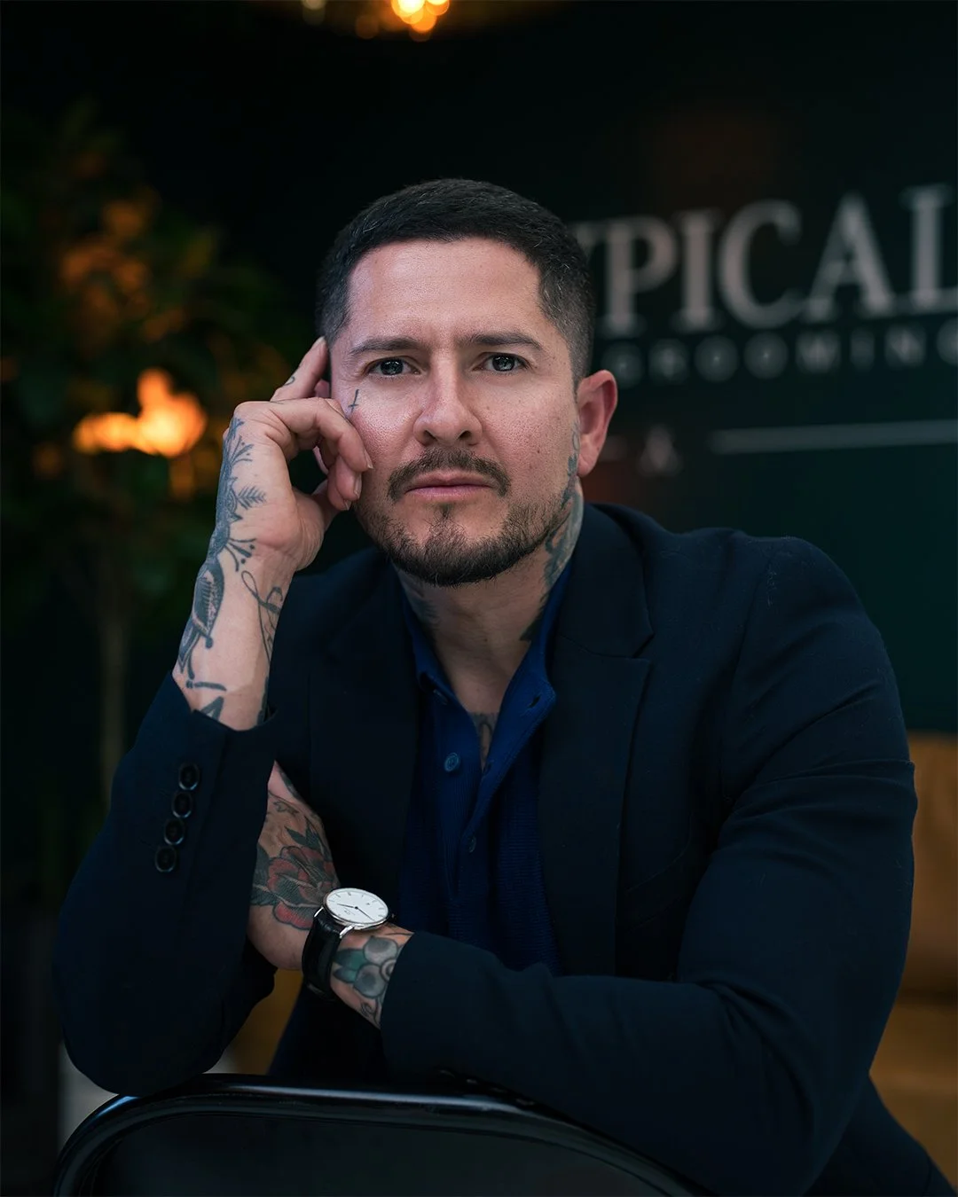 A man with short dark hair and tattoos on his arms and face, wearing a dark blazer and a blue shirt, sitting with his head resting on his hand in an indoor setting with blurred background and warm lighting.