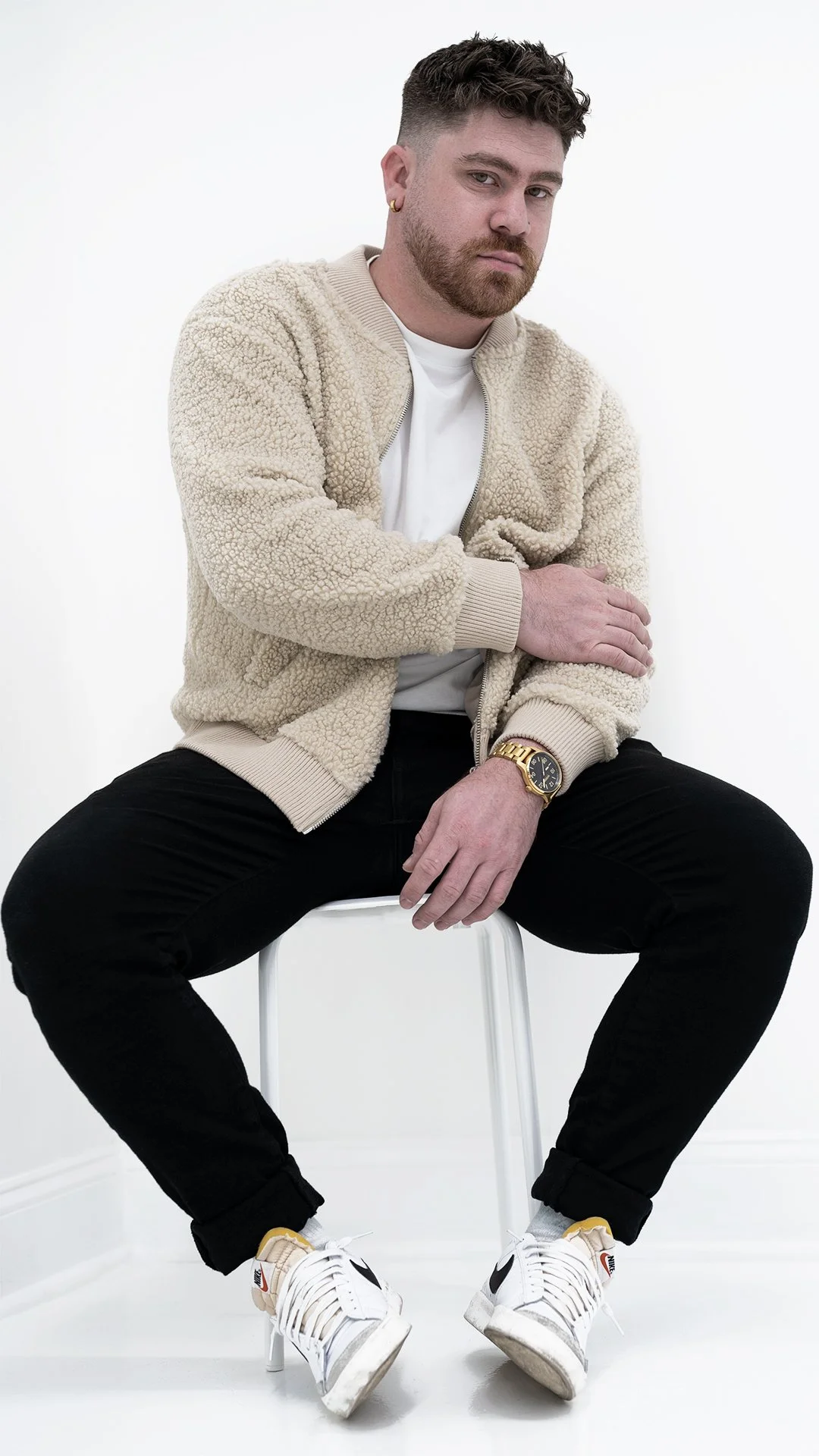 A man sitting on a white stool, wearing a beige sherpa zip-up jacket, white t-shirt, black pants, white sneakers, a gold watch, and gold earring, against a plain white background.