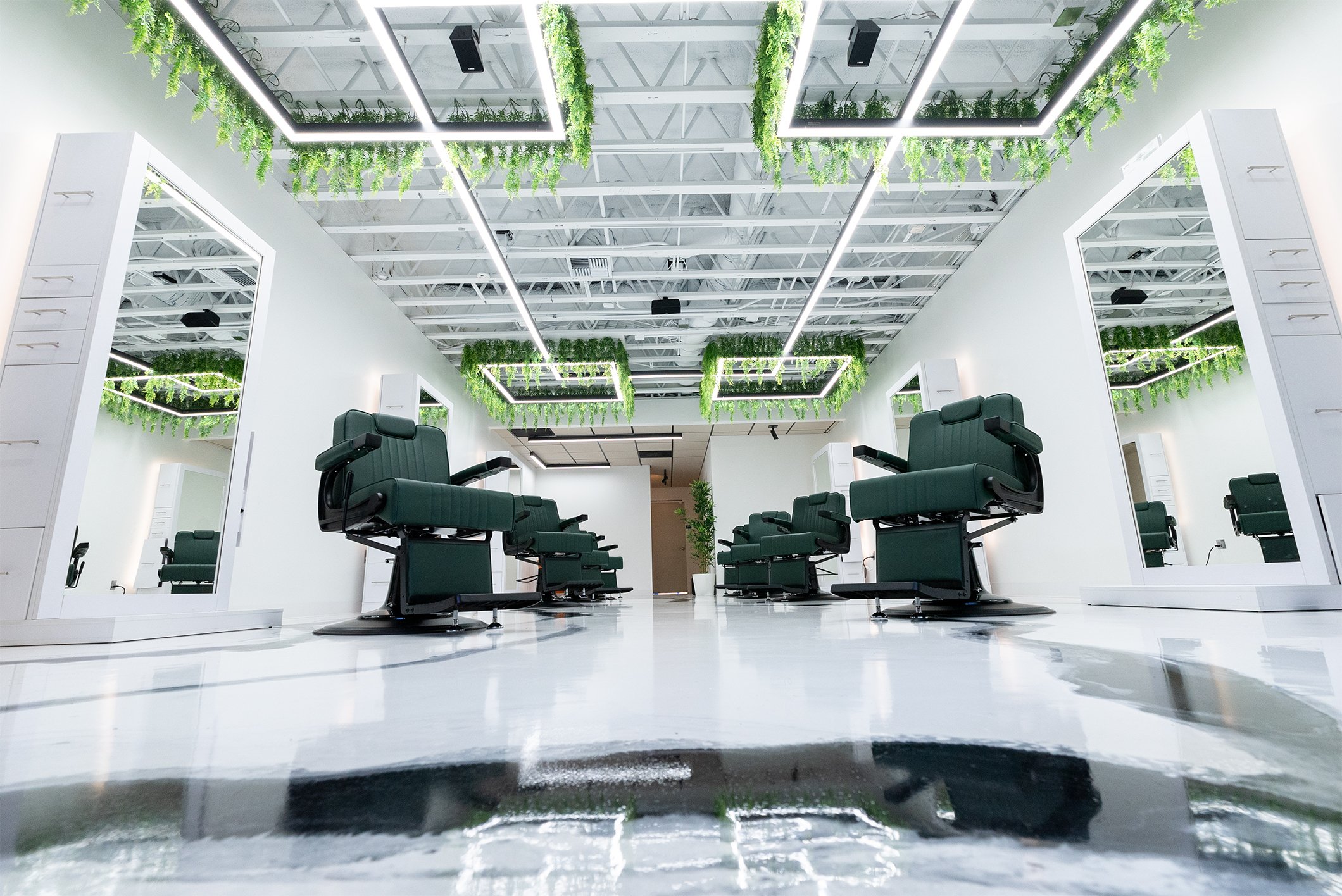Modern salon with barber chairs, white walls, mirrors, and ceiling decorated with hanging green plants and illuminated rectangular light frames.