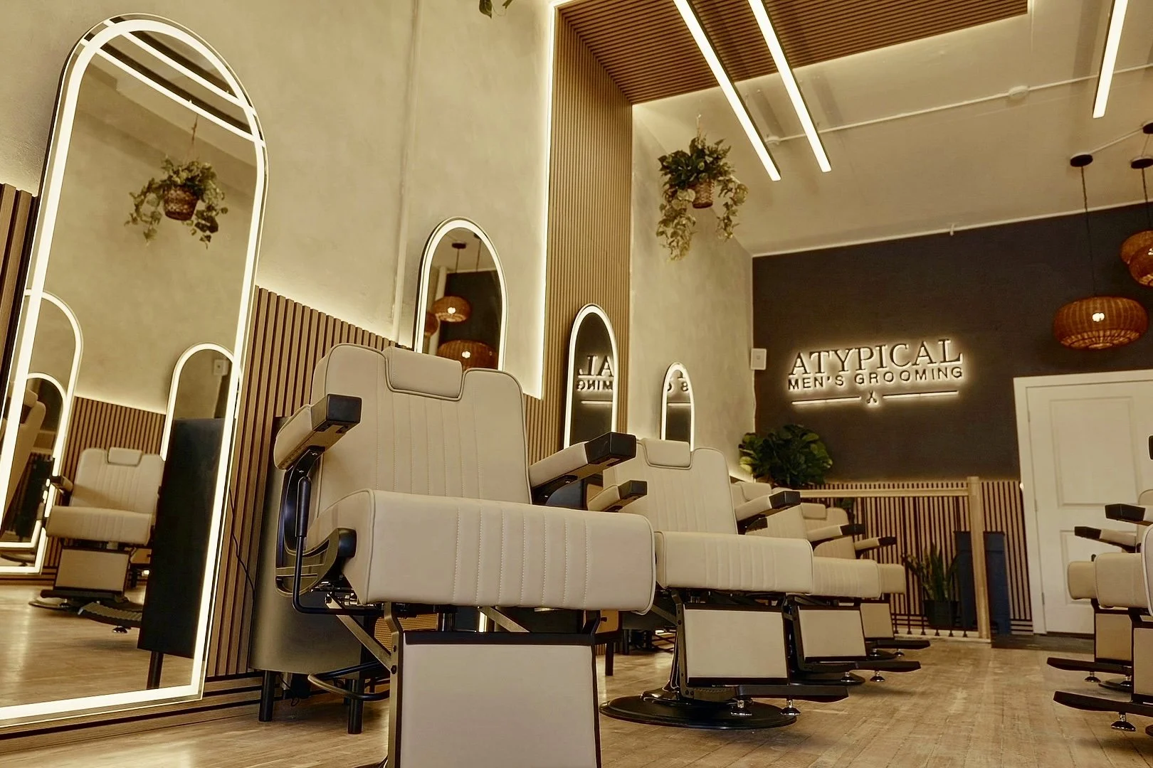 Interior of a men's grooming salon called 'Atypical Men's Grooming,' featuring rows of beige barber chairs, large illuminated mirrors, decorative hanging plants, and a dark accent wall with the salon name illuminated.