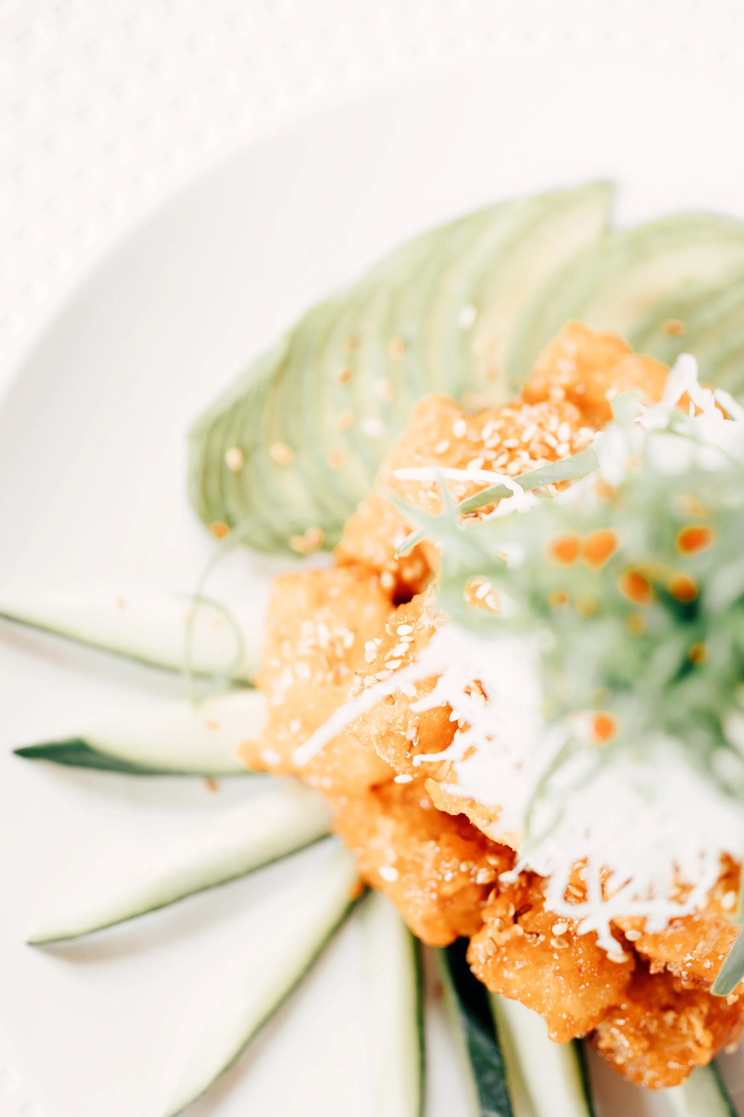 Tallerken med stekte biter av mat toppet med sesamfrø, avokado og agurkskiver, pyntet med grønnsaker.