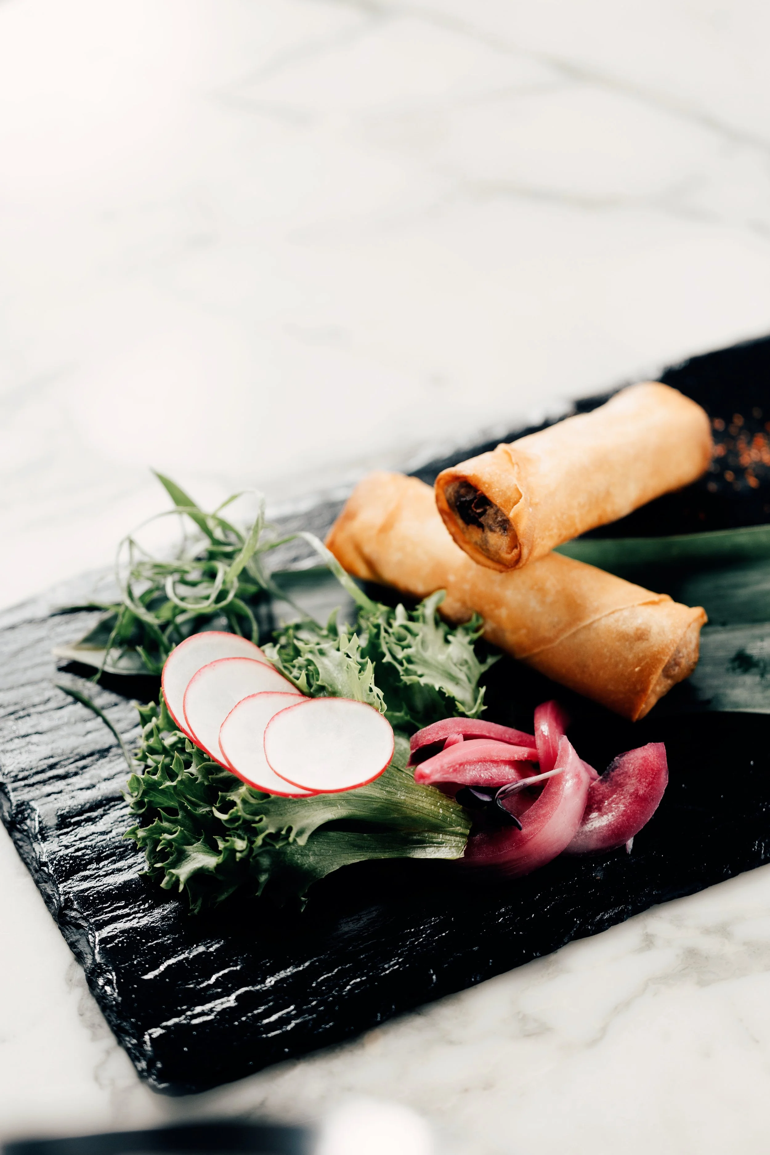 Vårruller med salat, skivet reddik, og syltet rødløk på svart skiferplate.