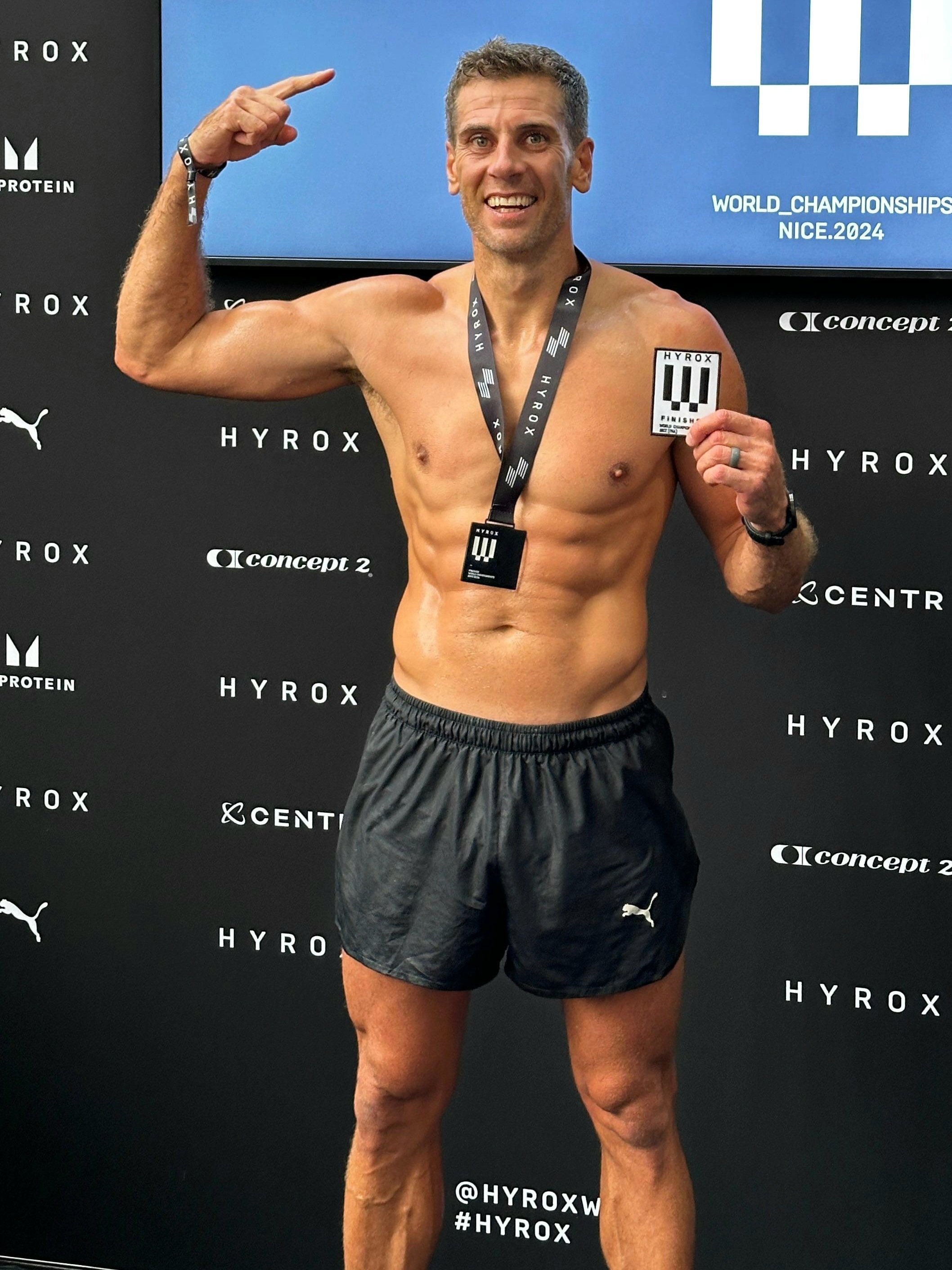 A shirtless man wearing black athletic shorts and a medal around his neck, smiling and holding a finishers' card, standing in front of a backdrop with logos for Hyrox and other sponsors, celebrating at a competitive event.