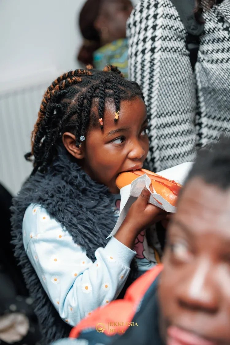A young girl with braided hair and earrings is eating a hot dog wrapped in paper at a gathering or party, surrounded by other people.