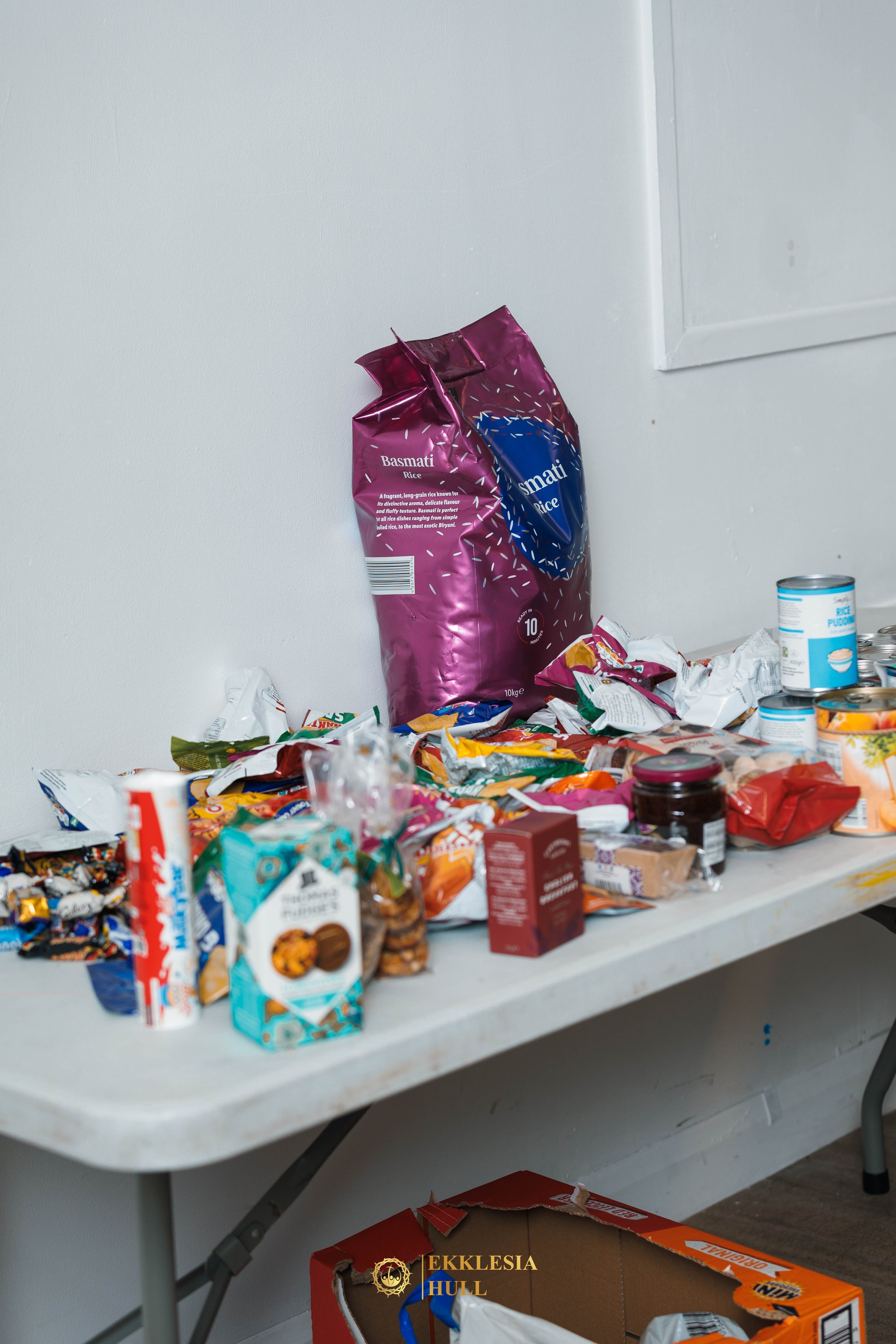 A cluttered table with various snack and food packages, including a large purple rice bag, cereal boxes, canned goods, and snack wrappers, in a room with a plain white wall and a cardboard box underneath.
