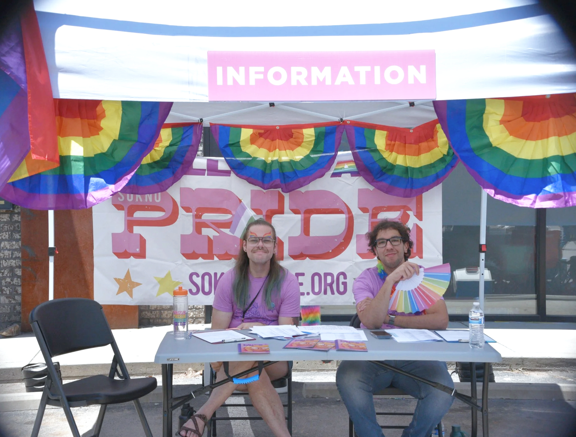 Two smiling SoKno Pride volunteers welcome visitors beneath colorful rainbow banners at the festival information booth.