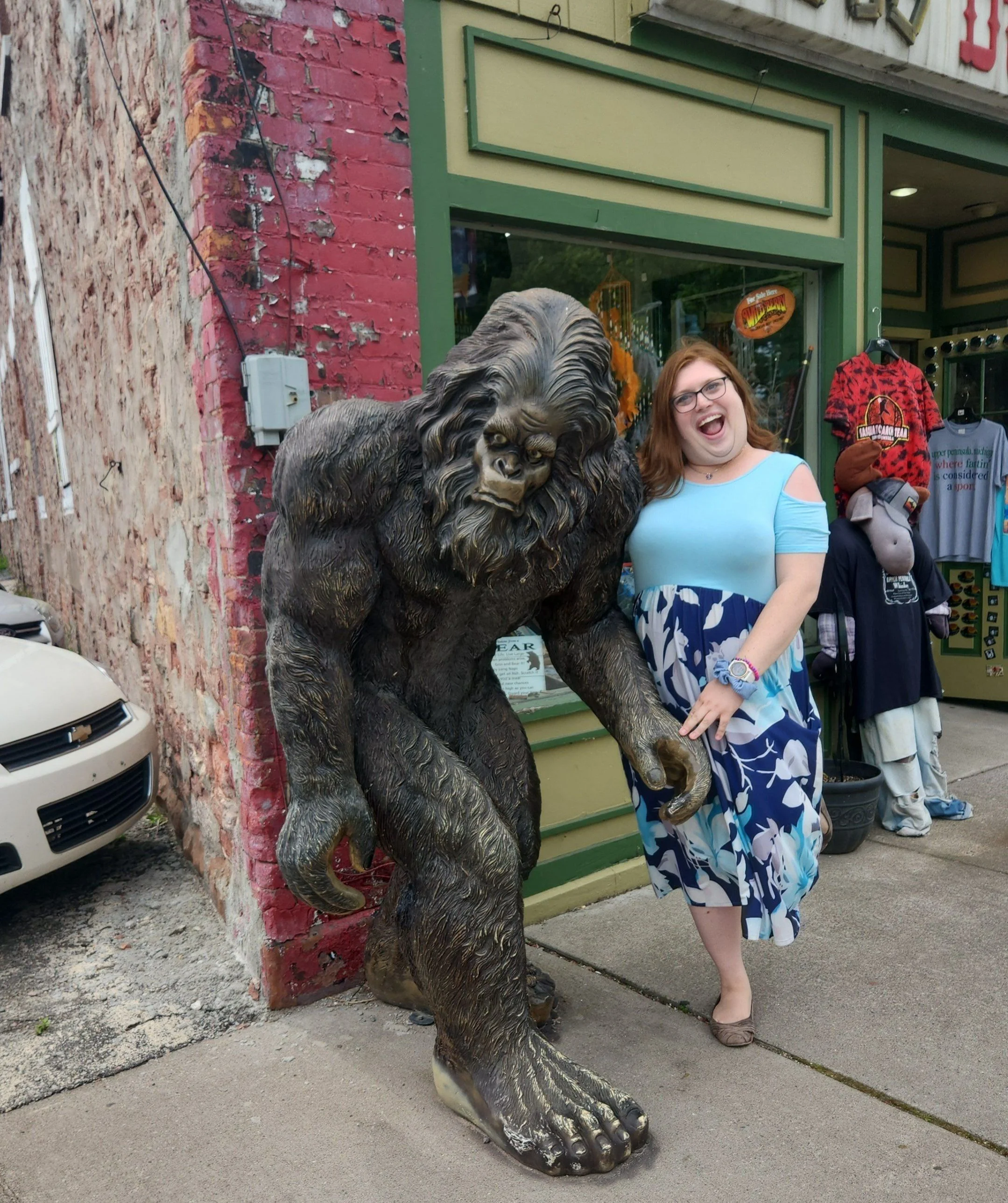 A woman with glasses and a blue and white dress smiling and pointing at a large realistic sculpture of a bear's paw next to her outside a store with a red and black patterned shirt on display.
