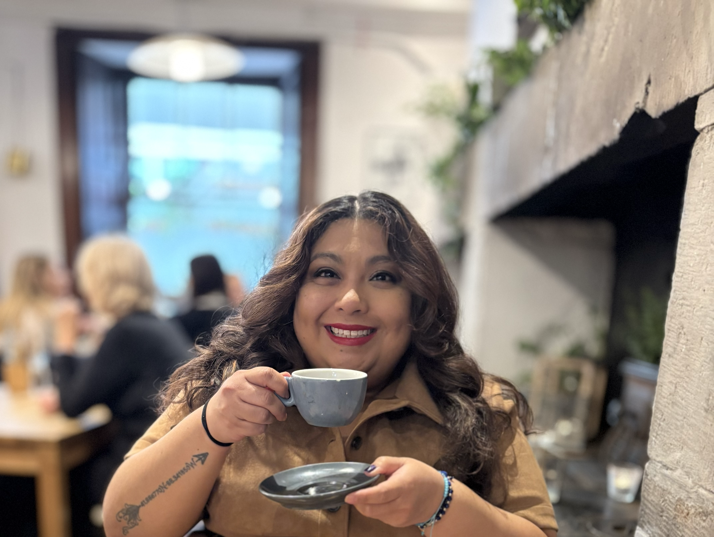 A woman with wavy brown hair and a tattoo on her left arm holds a mug and smiles at the camera. She is in a cozy indoor space with other people in the background, and a window with blue sky outside.