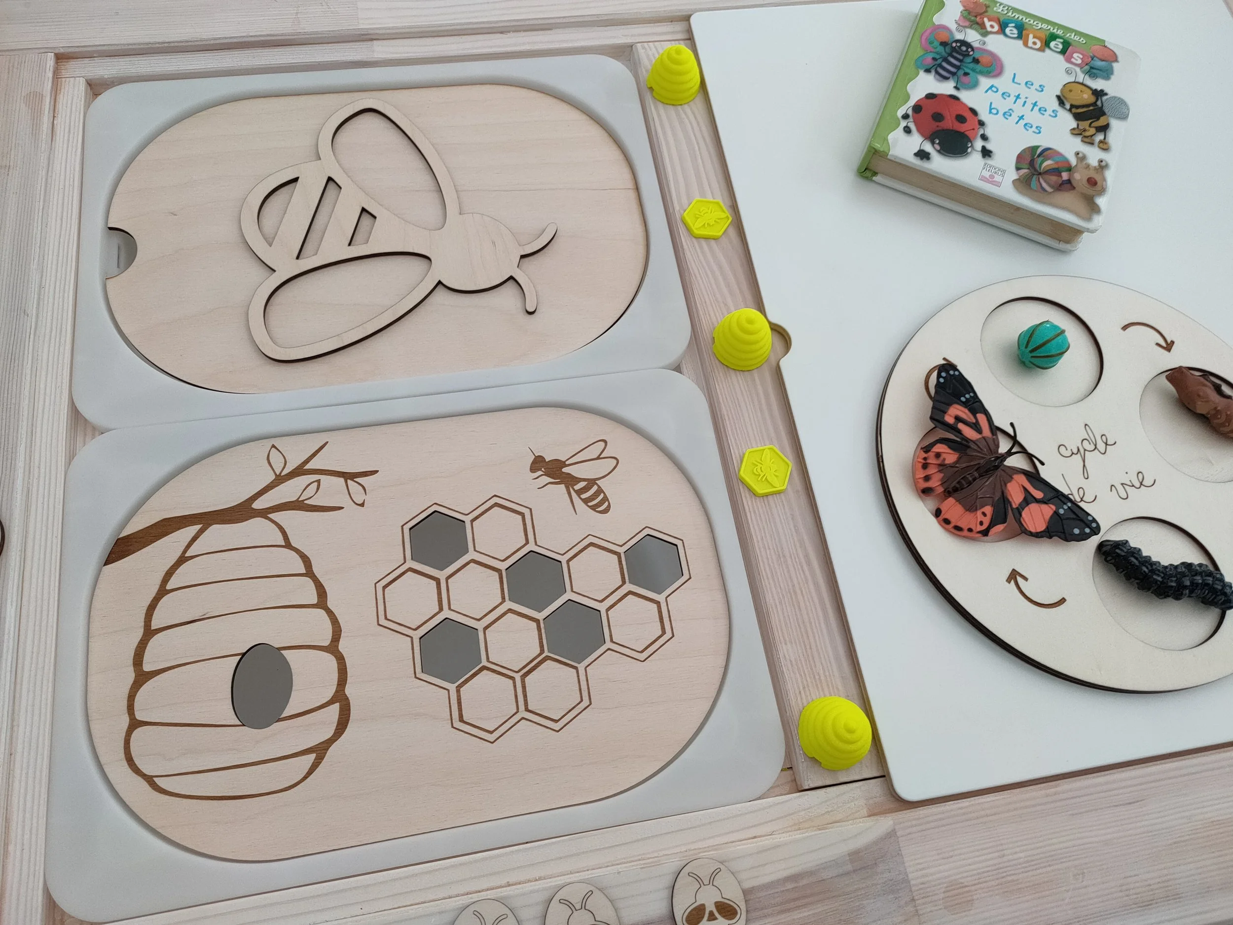 Un jeu éducatif avec des pièces en bois représentant des abeilles, des hyénomètres, des insectes, une ruche, un livre pour enfants avec des images d'insectes, une roue de vie et des pièces en forme d'insectes.