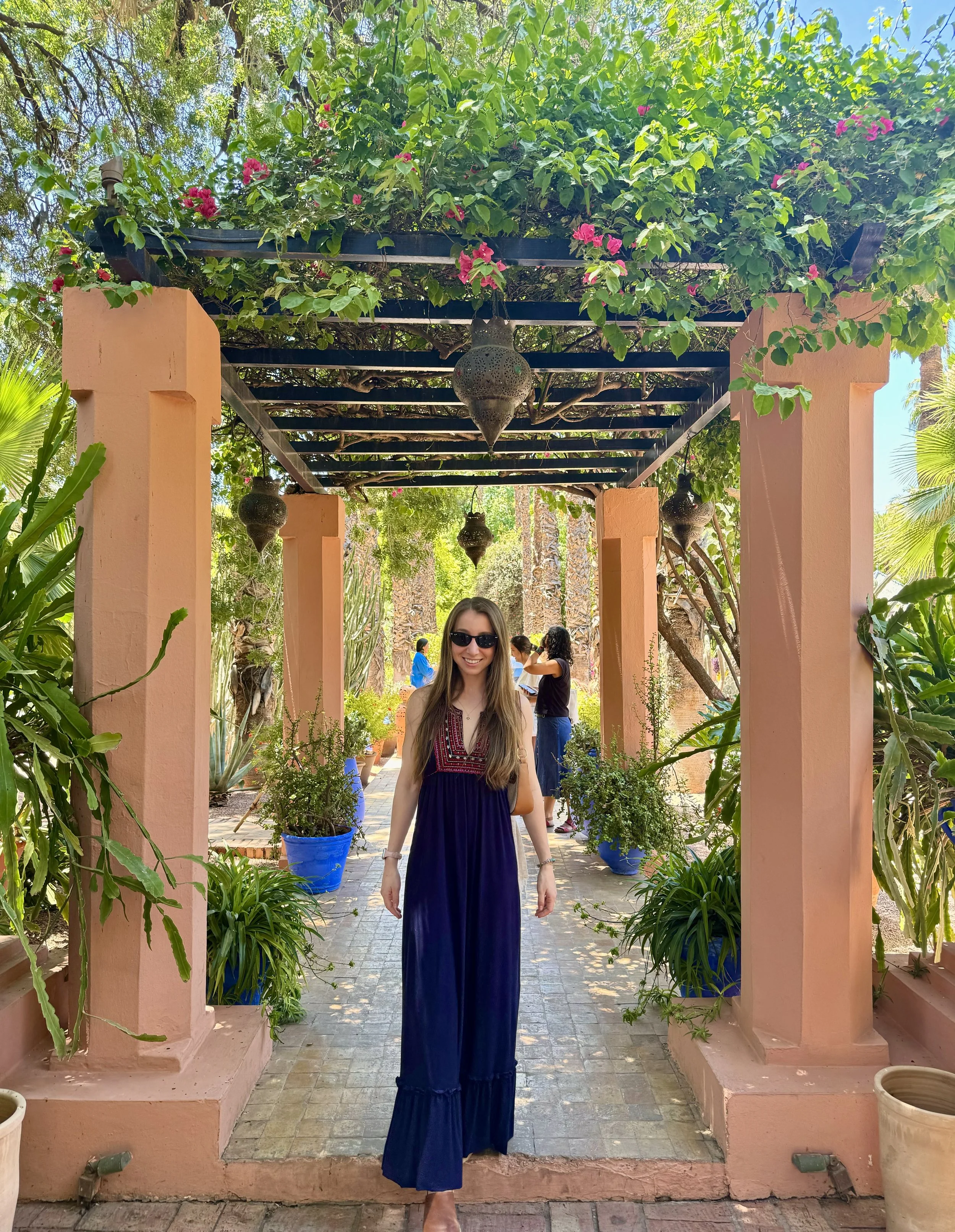A woman wearing sunglasses and a long blue dress walking through a garden arched walkway with potted plants and hanging lanterns, lush green plants, and other people in the background.