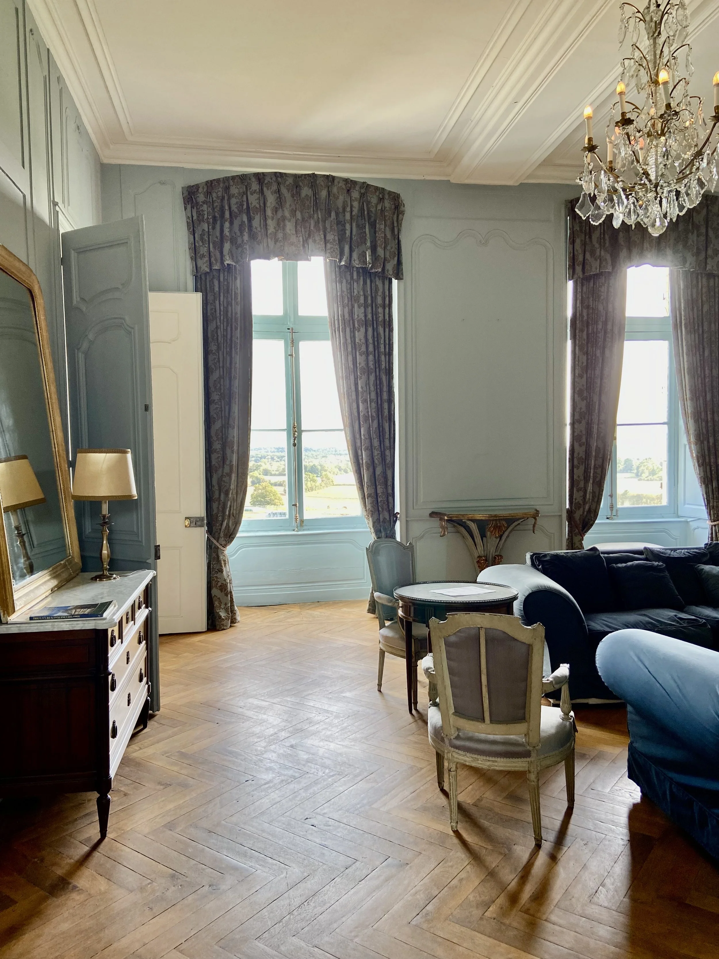 Elegant living room with vintage furniture, wooden herringbone floor, large windows with patterned curtains, a chandelier, and a mirror on a cabinet.