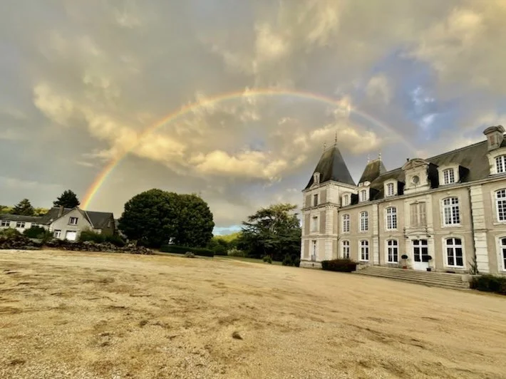 Château-style building with a rainbow in the sky, surrounded by trees and a gravel courtyard.
