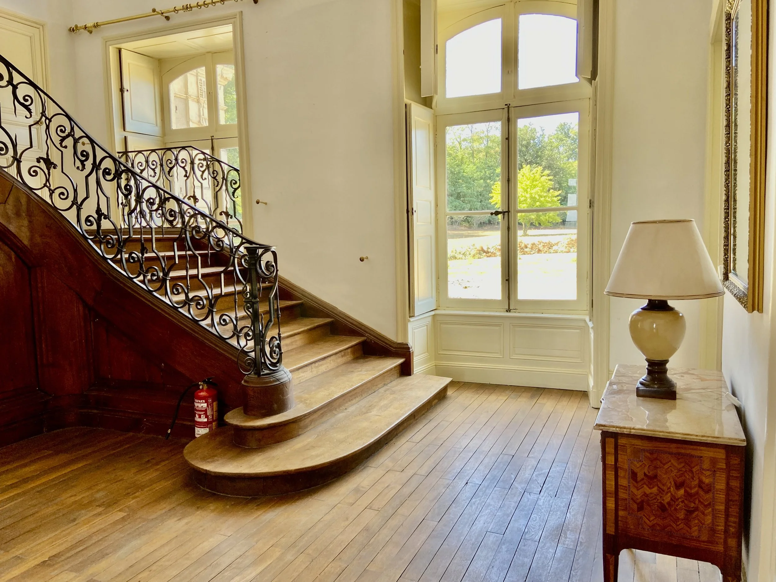 Elegant interior with a wooden staircase, ornate railing, large window, wooden flooring, small table with a lamp, and a fire extinguisher.