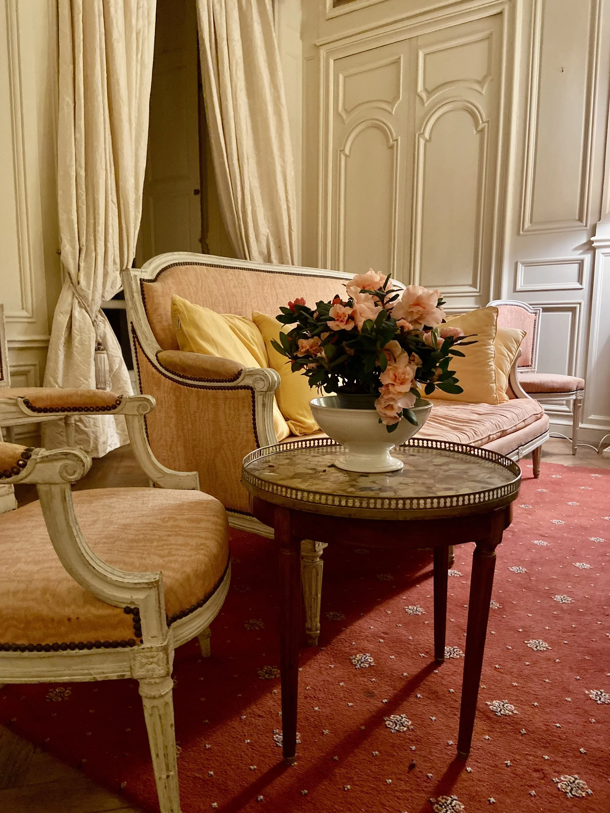 Elegant living room with vintage furniture, including an upholstered sofa, armchair, and a round side table with a floral centerpiece. Cream-colored curtains and paneling, with a patterned red carpet.