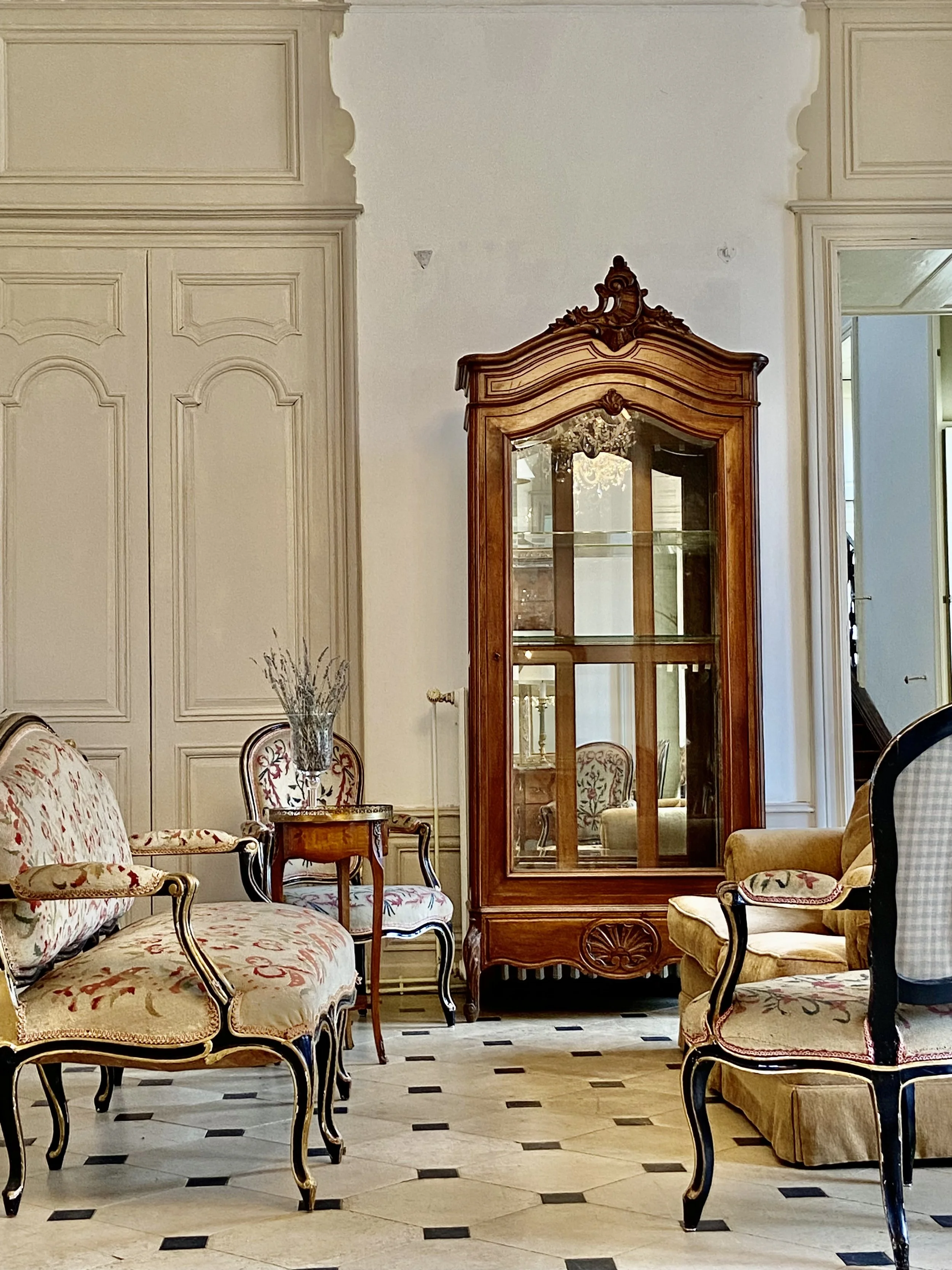 Elegant room with vintage furniture including a floral-upholstered sofa, a matching chair, an antique wooden cabinet with glass doors, and a small side table, all set against a paneled wall with a tiled floor.