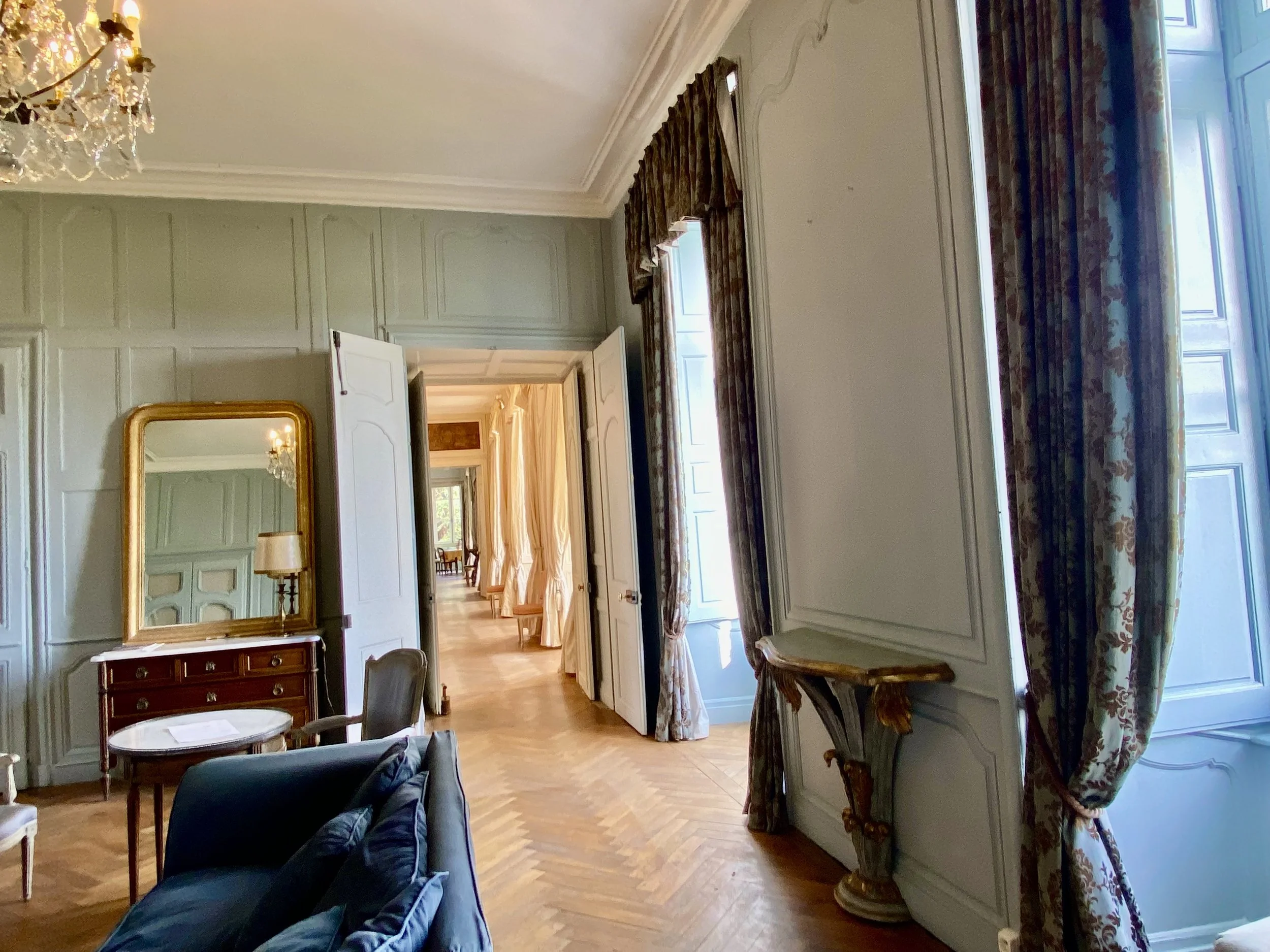 Elegant room with a chandelier, vintage furniture, large windows with floral curtains, and a decorative mirror on the wall.