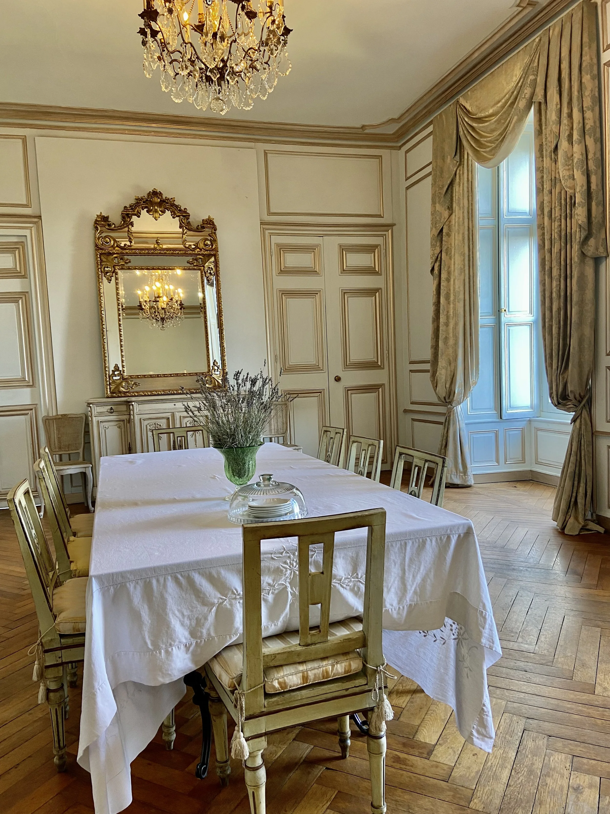 Elegant dining room with a long table covered by a white tablecloth, surrounded by chairs. A decorative glass dish and a vase with flowers are on the table. The room features a chandelier, ornate mirror, and large windows with curtains, showcasing cl