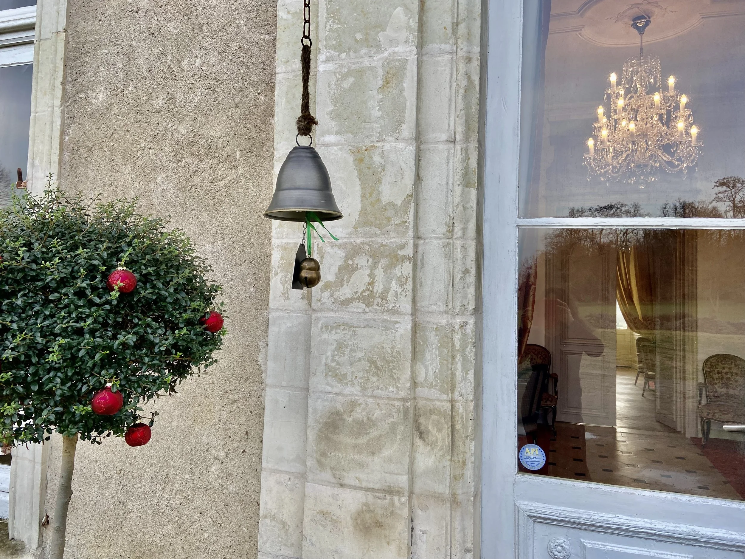 Exterior view of a building with a hanging bell, ornamental shrub with red decorations, and a glass door reflecting a chandelier and indoor furniture.