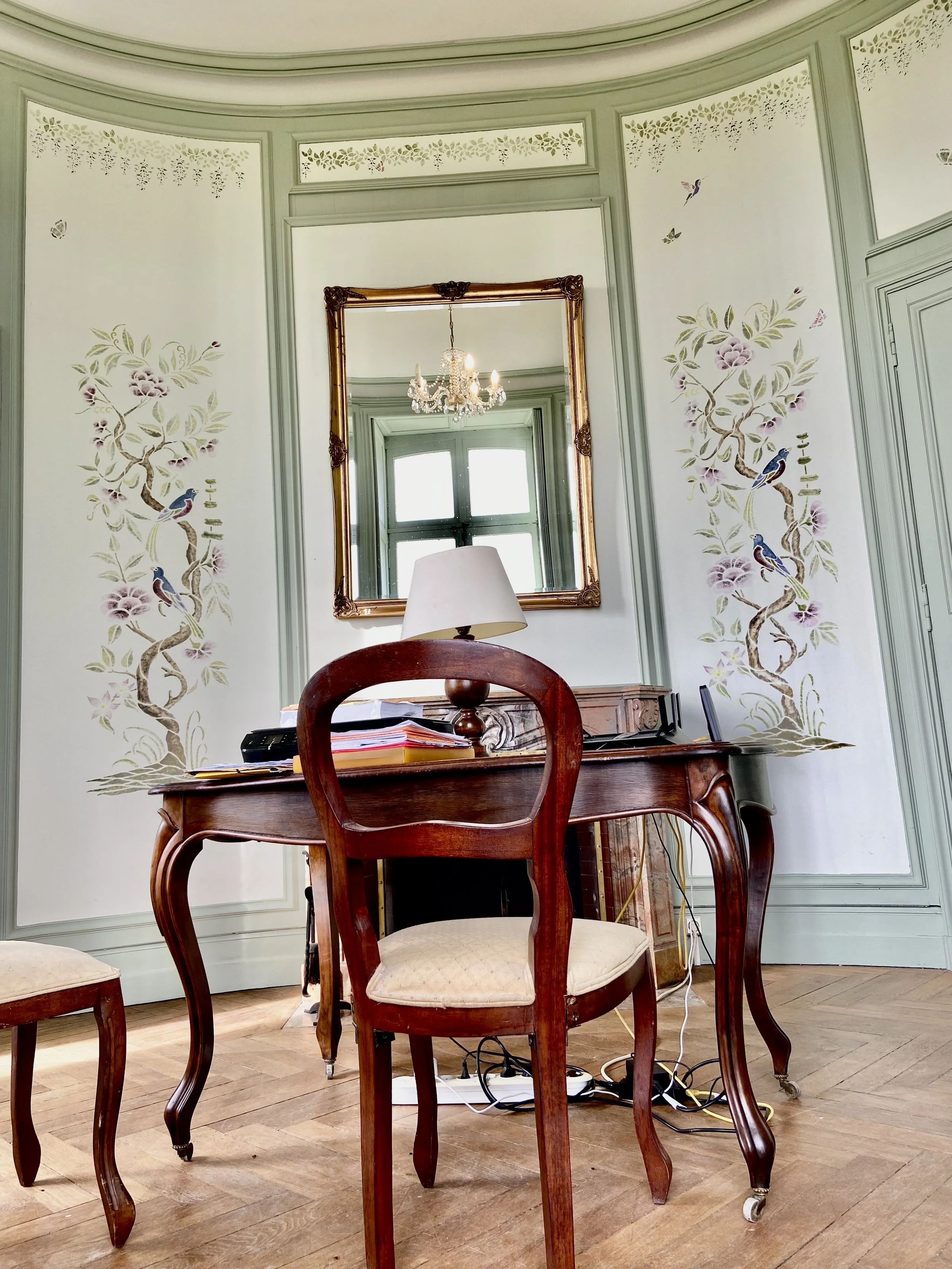 Elegant room with ornate green and white walls featuring floral and bird designs. A wooden desk and chairs with curved legs are in the foreground. A mirror on the wall reflects a chandelier and window. Light streaming in gives a cozy atmosphere.