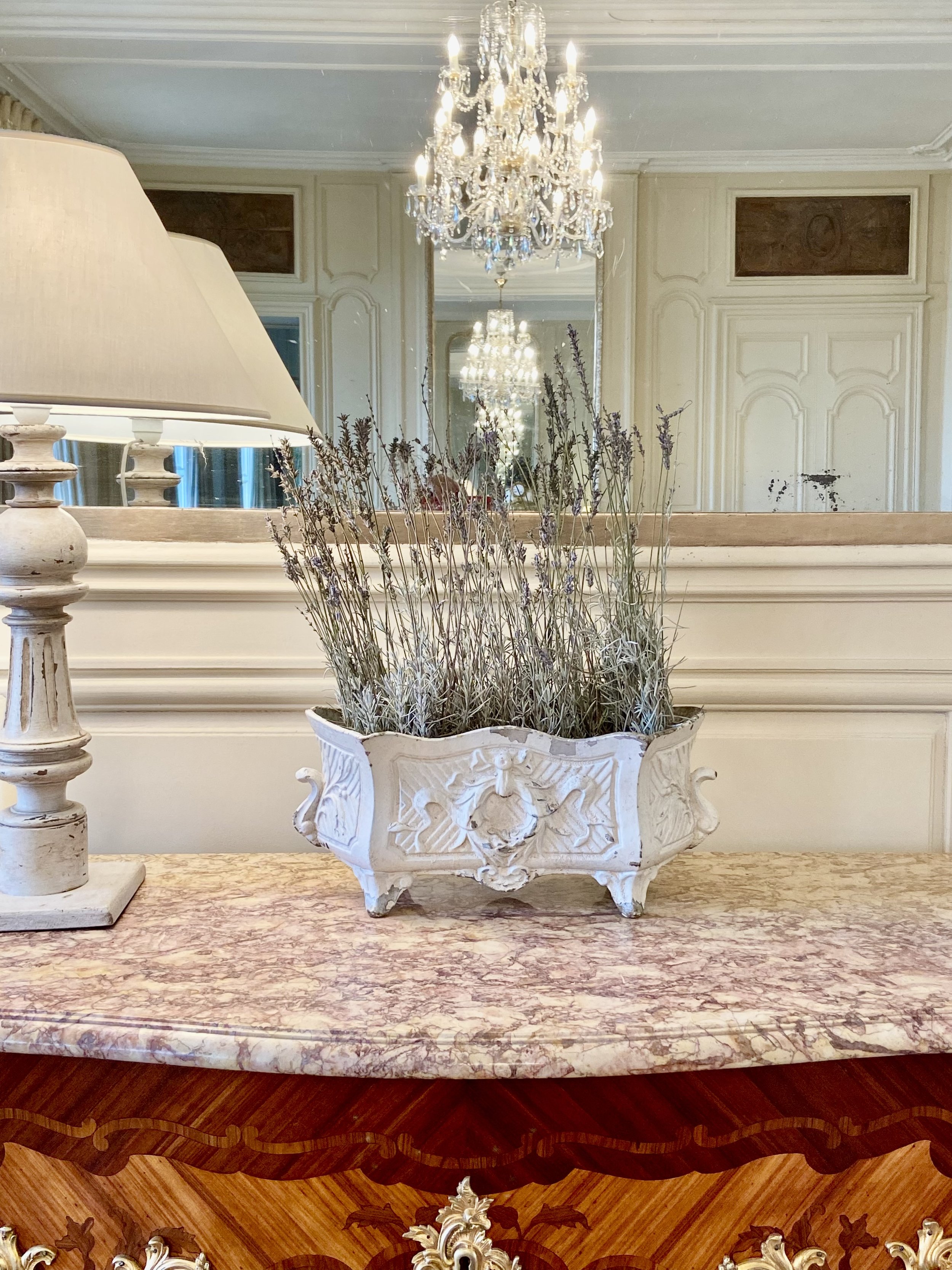 Elegant room with a chandelier, ornate mirror, and a marble-topped table featuring a vintage lamp and decorative vase with dried lavender.