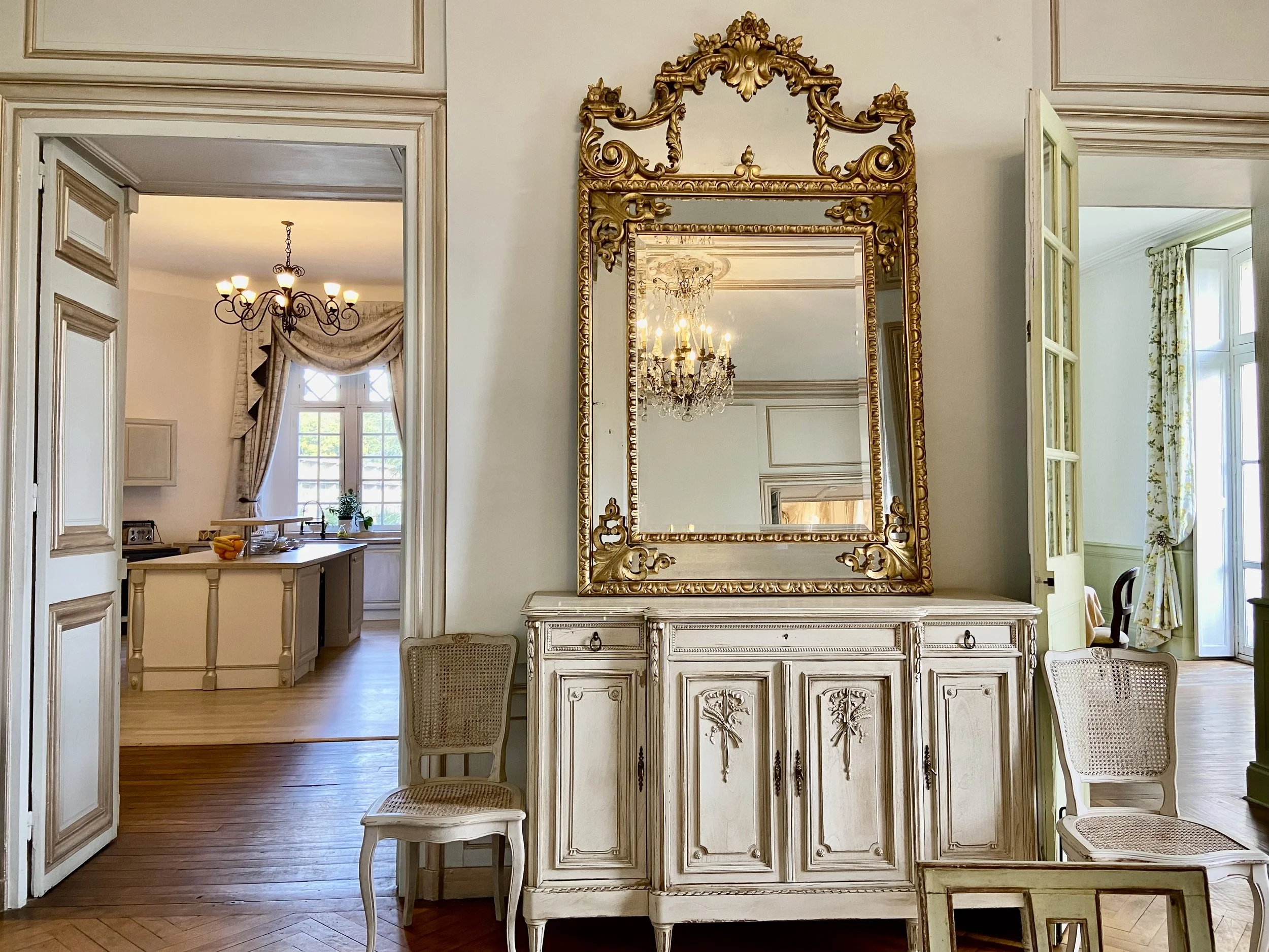 Vintage-style interior with an ornate mirror above a decorative cabinet, two white chairs, open doorways leading to a chandelier-lit room with a large window and draped curtains.