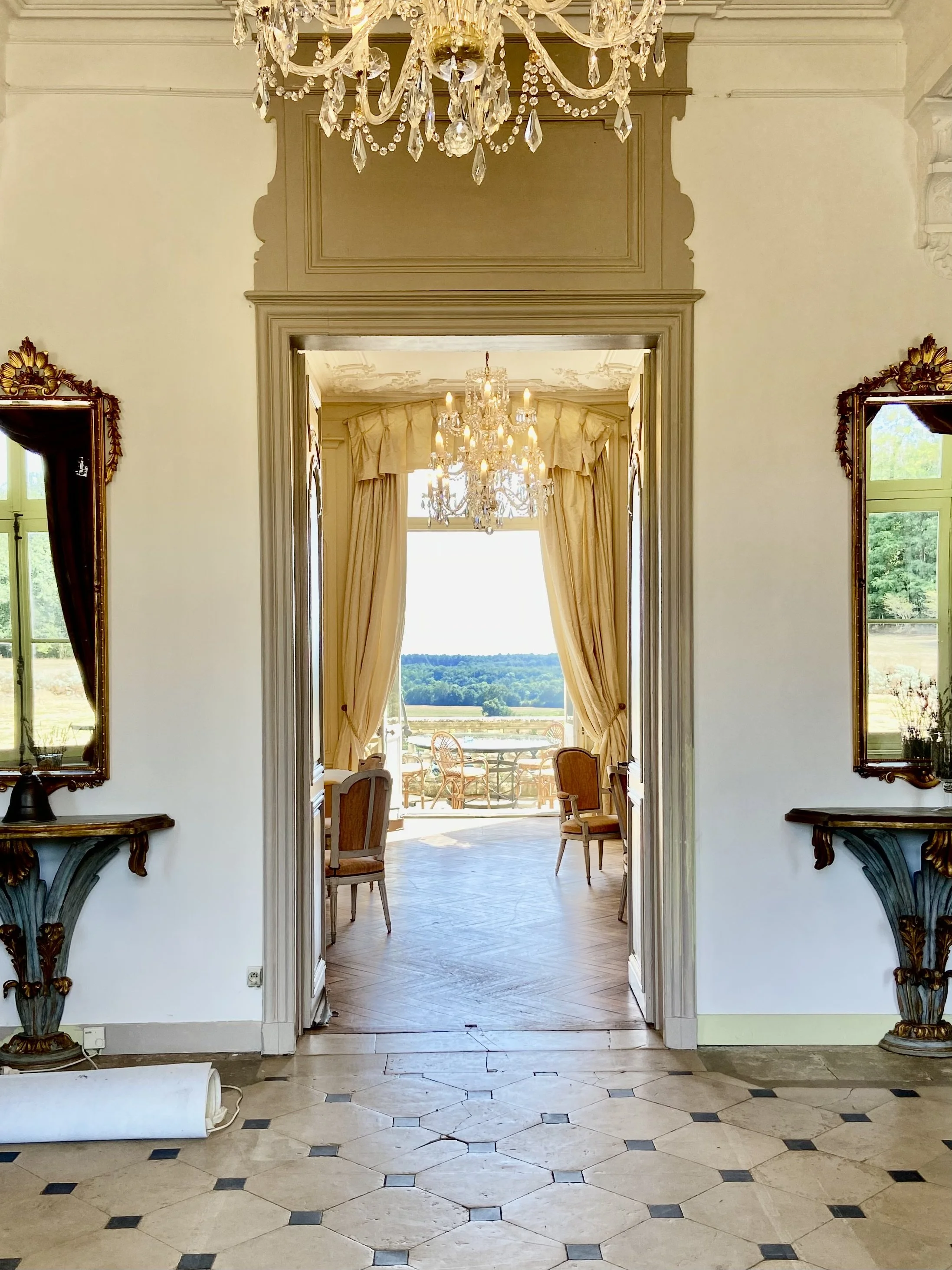Elegant interior with chandeliers, mirrors, decorative tables, and a view to an outdoor patio with chairs.
