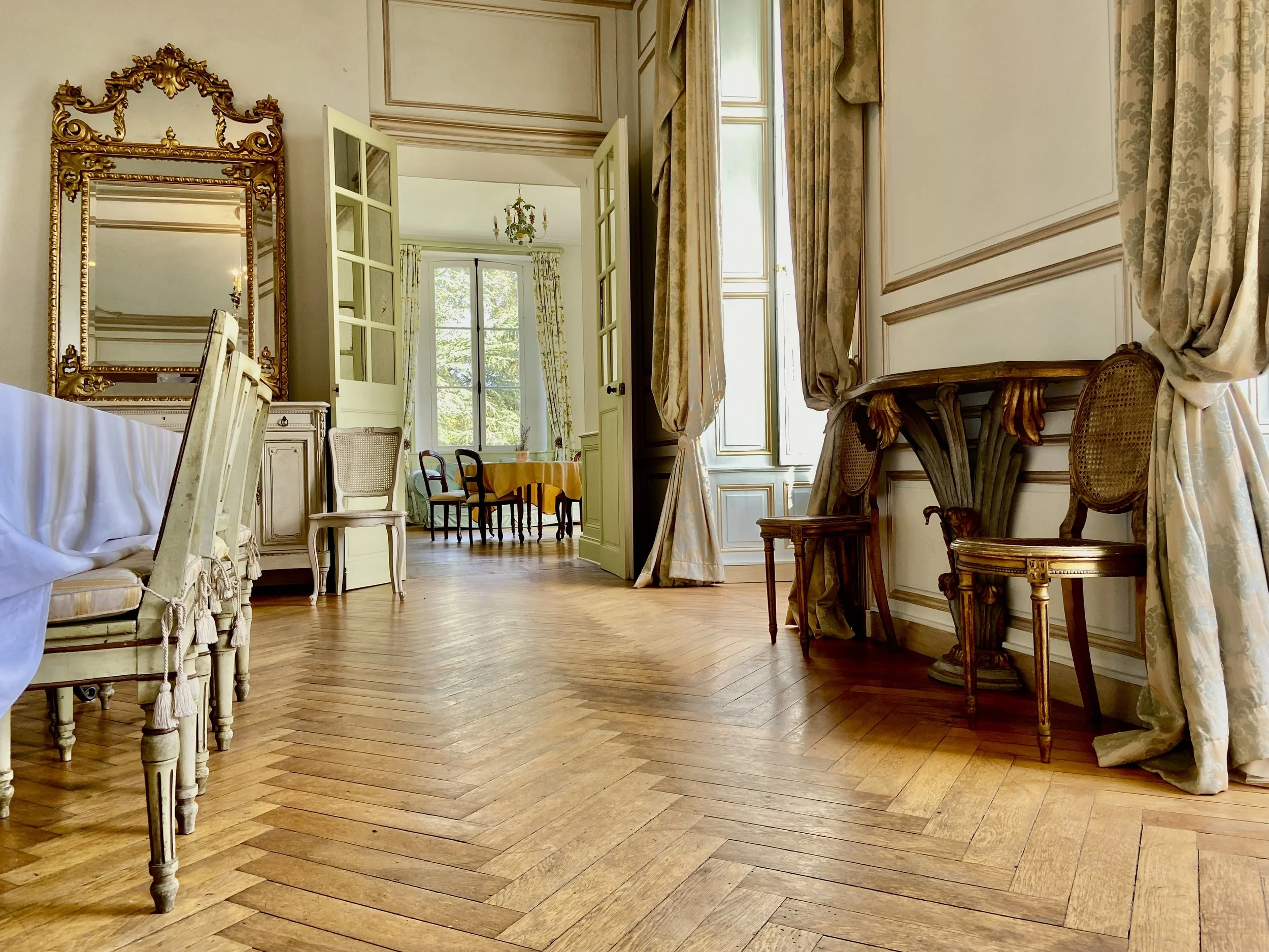 Elegant room with wooden floors, ornate mirror, and vintage furniture, featuring drapes and chairs, leading to a dining area in the background with a table and several chairs.