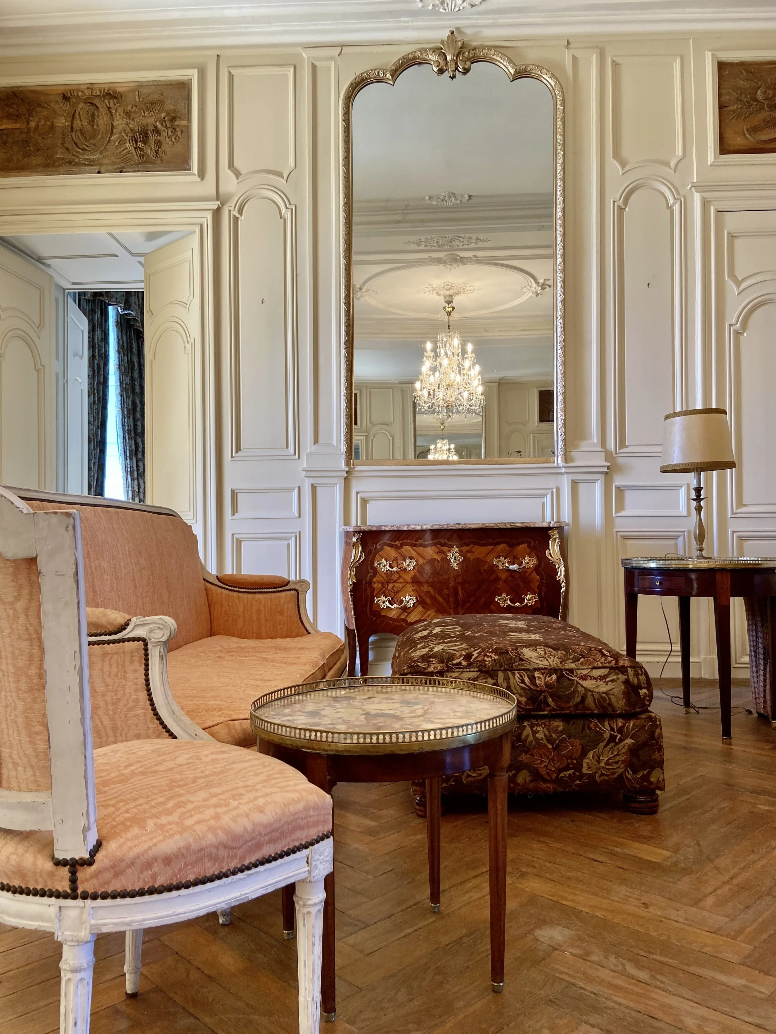 Elegant living room with antique furniture including pastel upholstered armchairs, a coffee table with a metallic rim, a large ornate mirror above a fireplace, and a chandelier reflection.