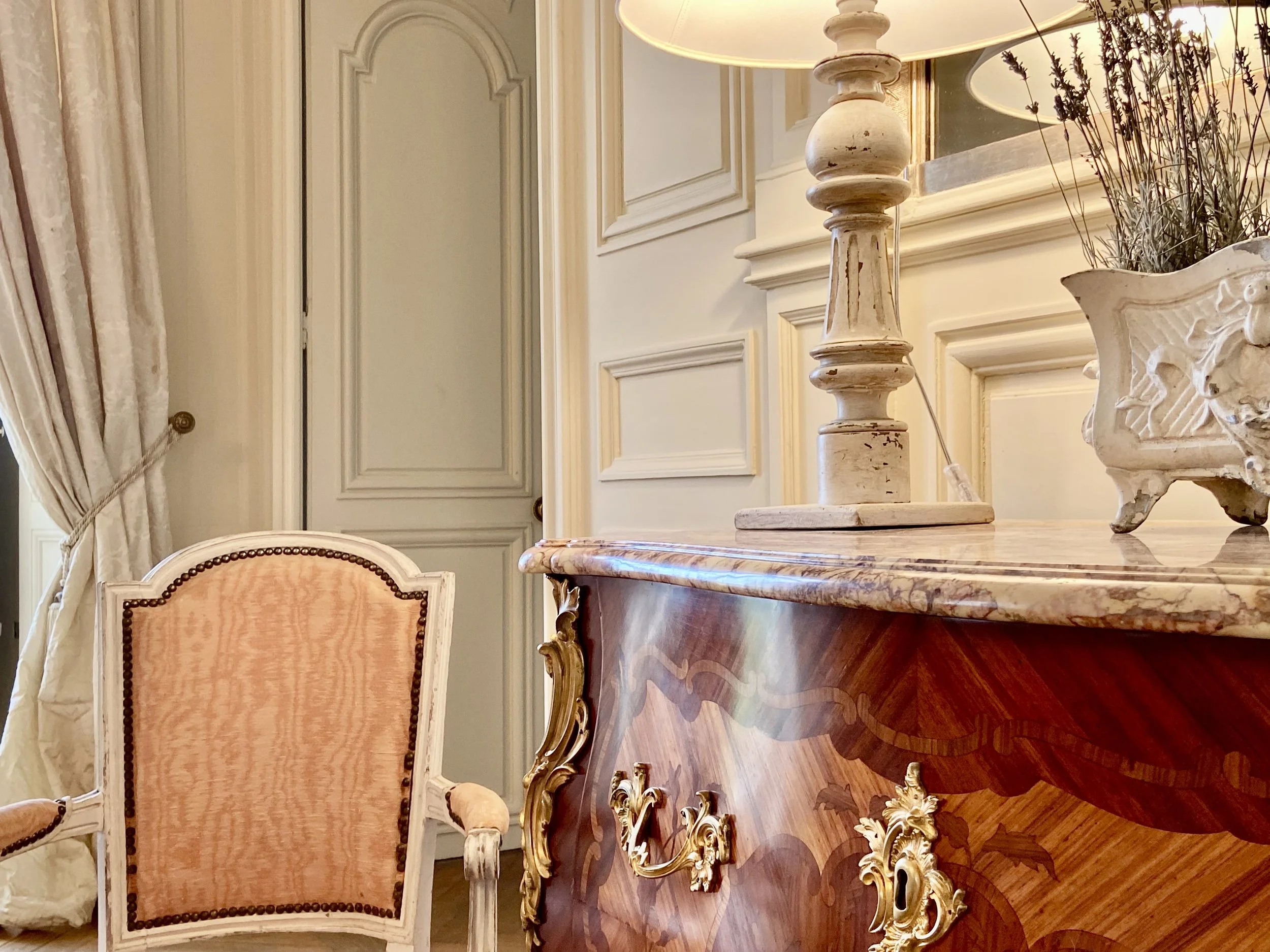 Elegant interior with vintage armchair, ornate wooden dresser with marble top, decorative lamp, and potted plant in a lavish room setting.