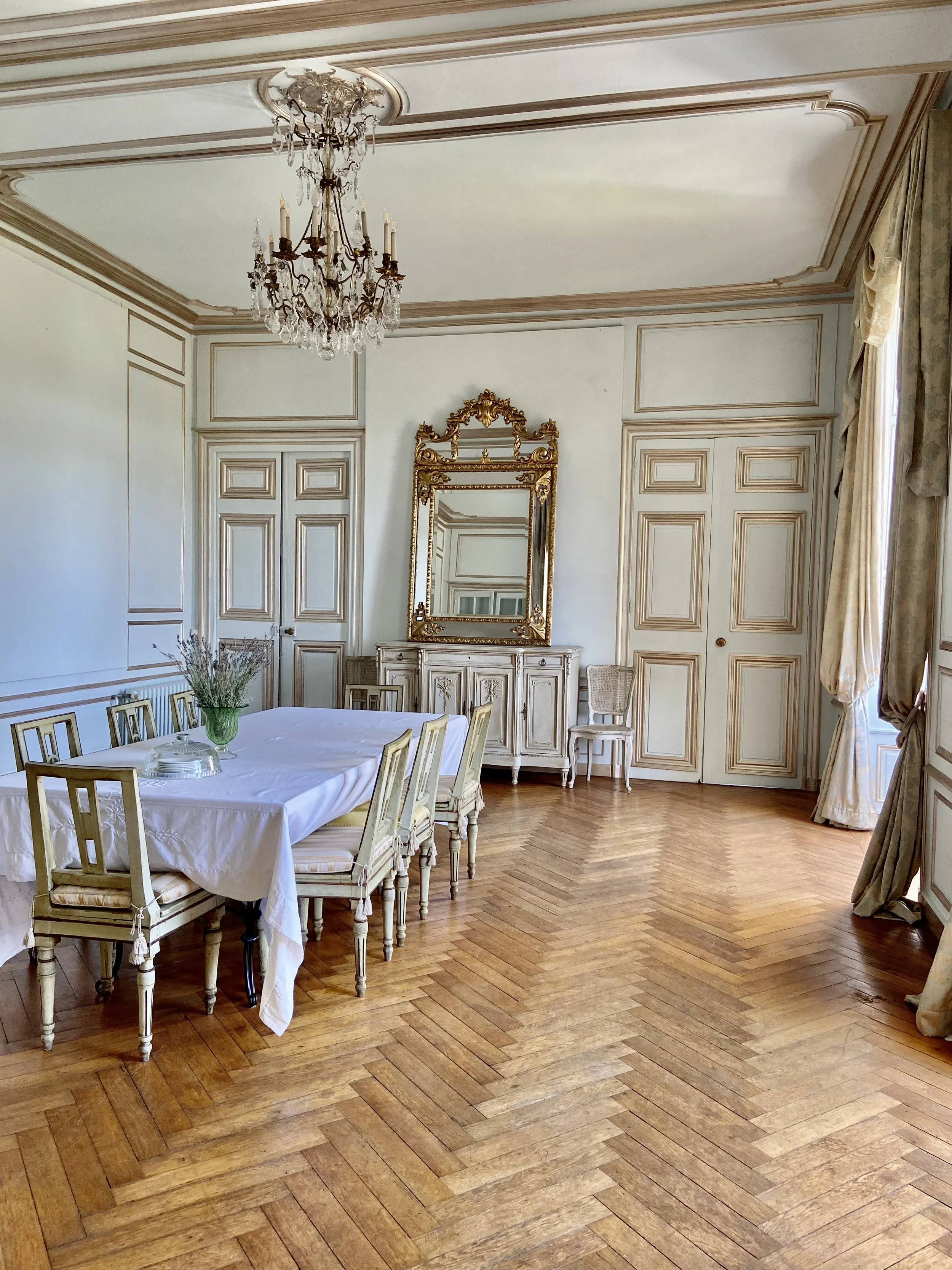 Elegant dining room with vintage decor, featuring a chandelier, large mirror, antique table with chairs, hardwood floors, and paneled walls.