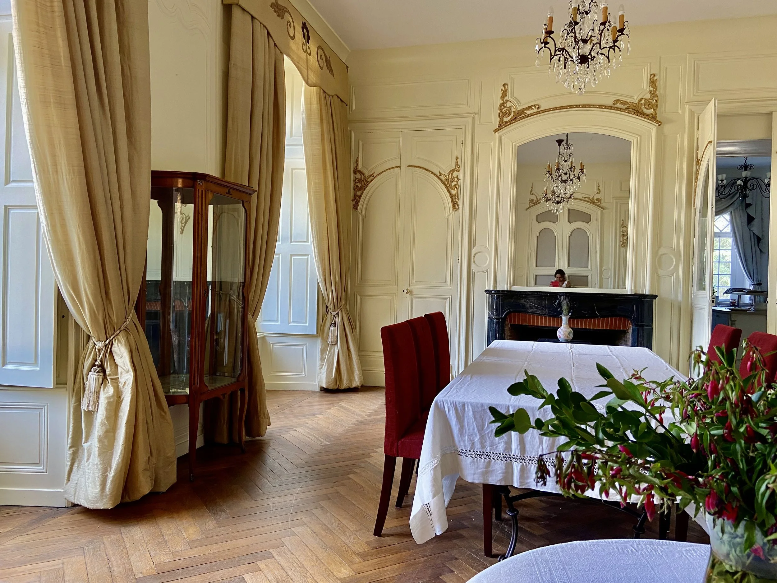 Elegant dining room with a long table covered in a white tablecloth, surrounded by red upholstered chairs, beige curtains, ornate moldings, a chandelier, and decorative plants.