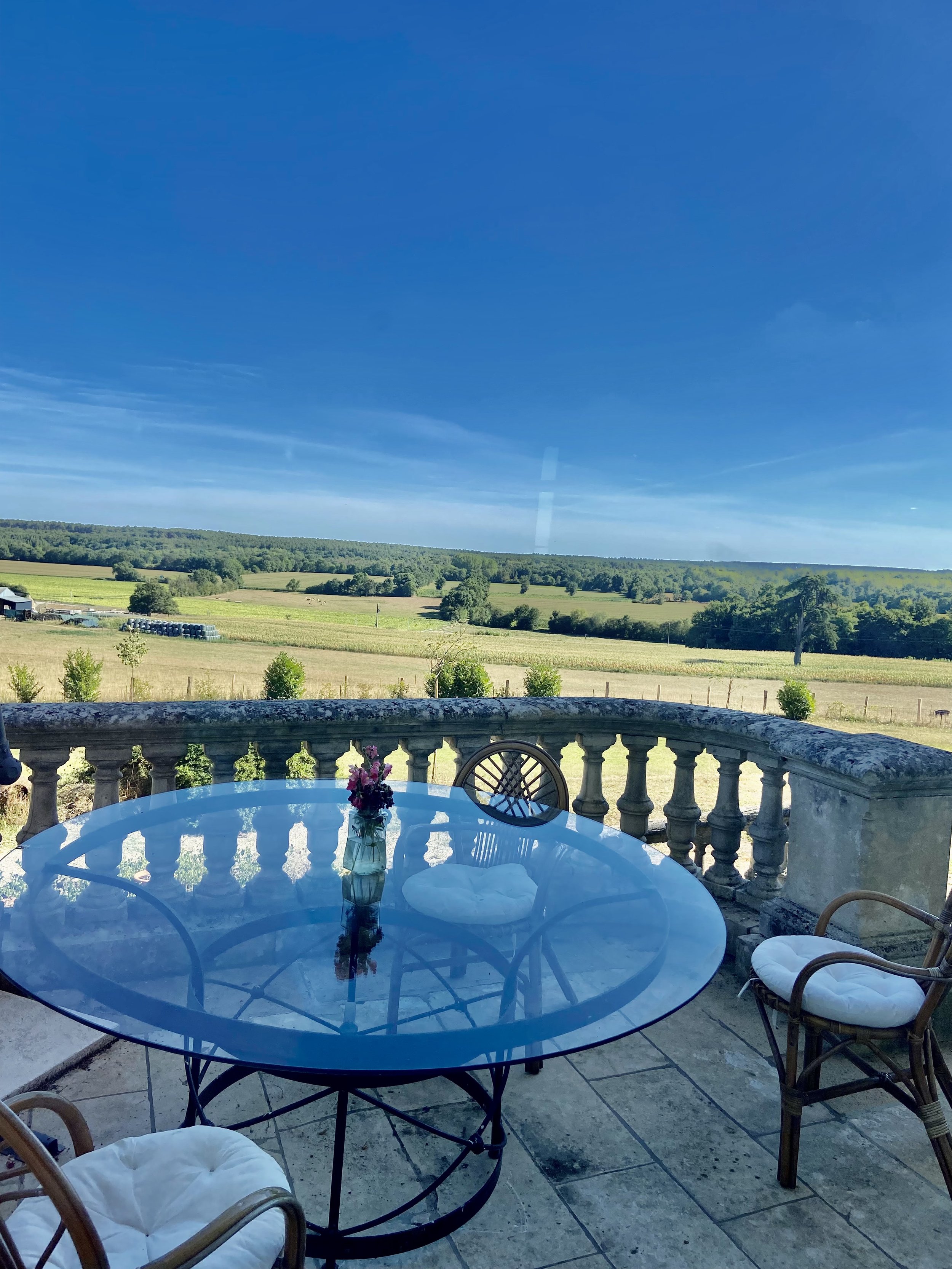 Glass table with chairs on a patio overlooking a scenic landscape with fields and trees under a clear blue sky.
