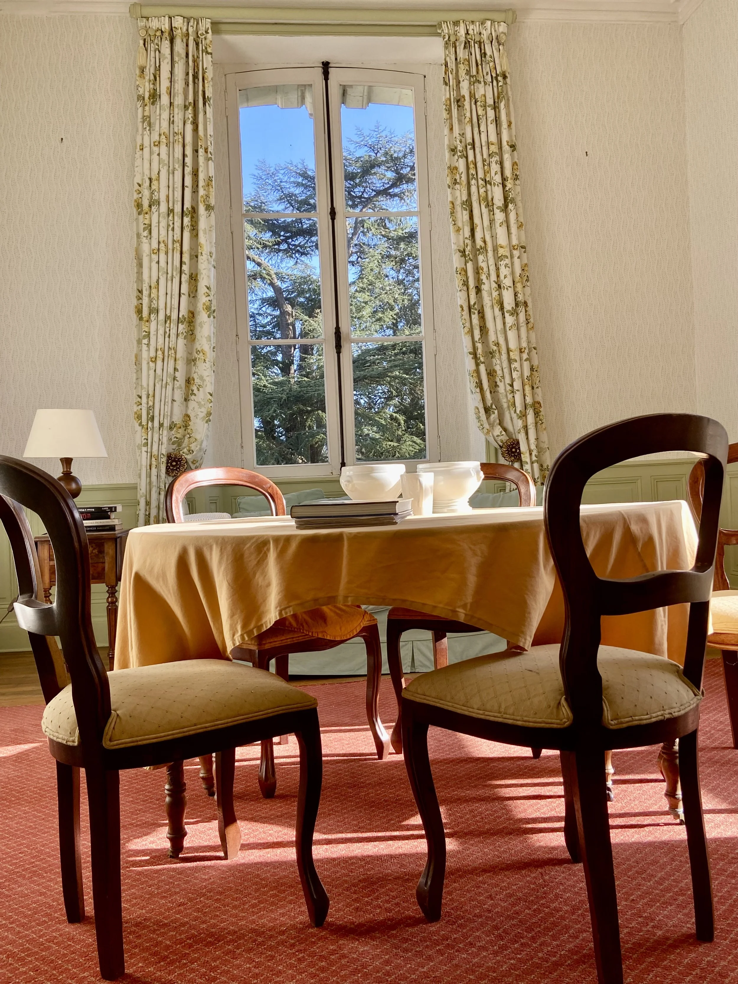 Dining room with a table covered by a yellow tablecloth, four chairs, a lamp, and a window with floral curtains overlooking trees.
