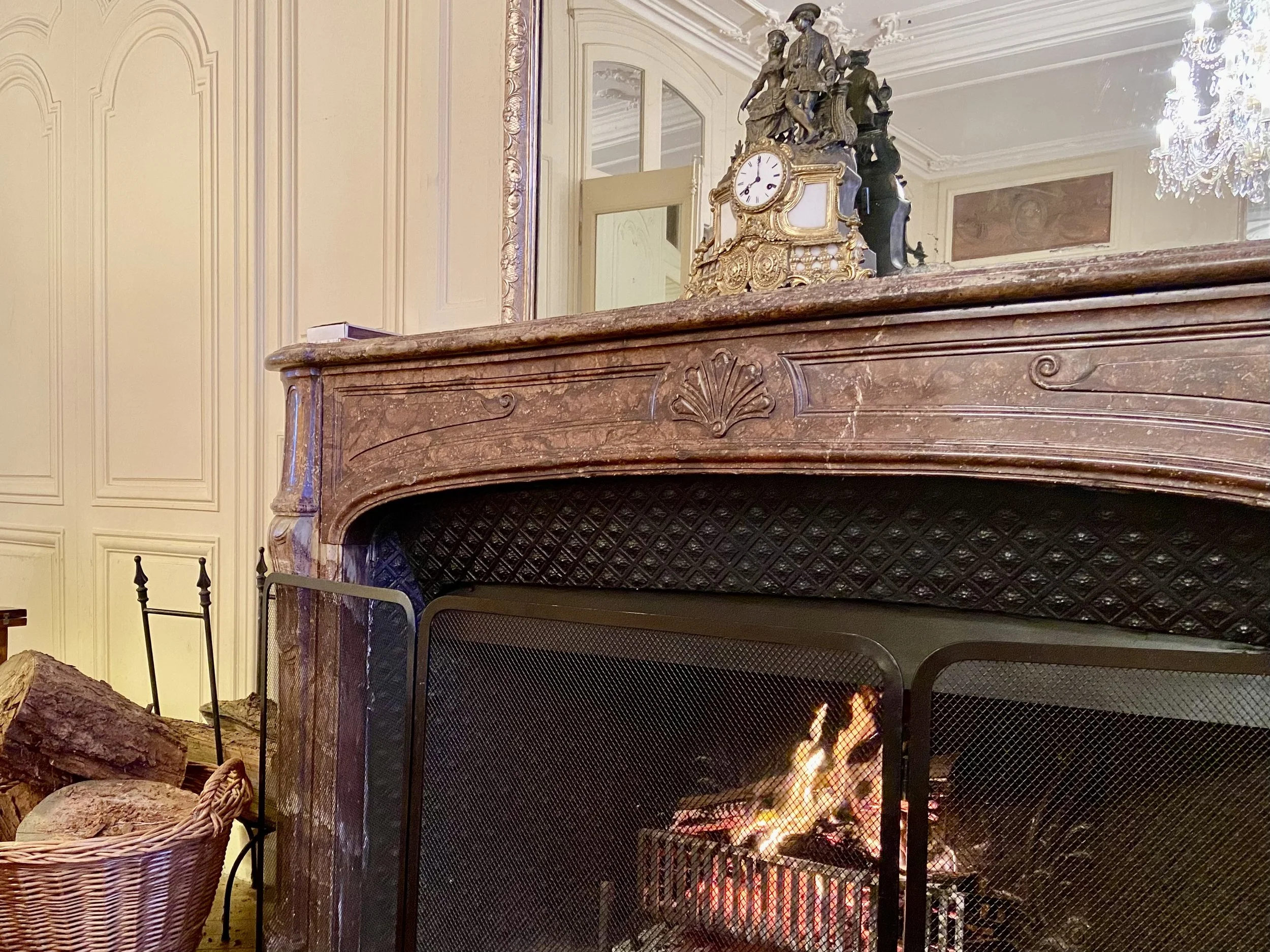 A traditional fireplace with a lit fire and a decorative mantelpiece, including a clock and figurines, in an ornate room with wooden paneling and a chandelier.