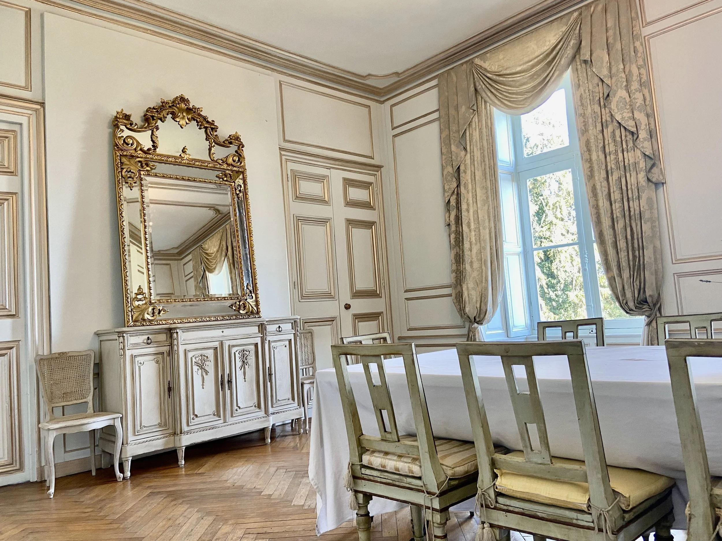 Elegant dining room with antique furniture, featuring a long table, vintage chairs, a large ornate mirror on a decorative sideboard, and luxurious curtains framing a window.